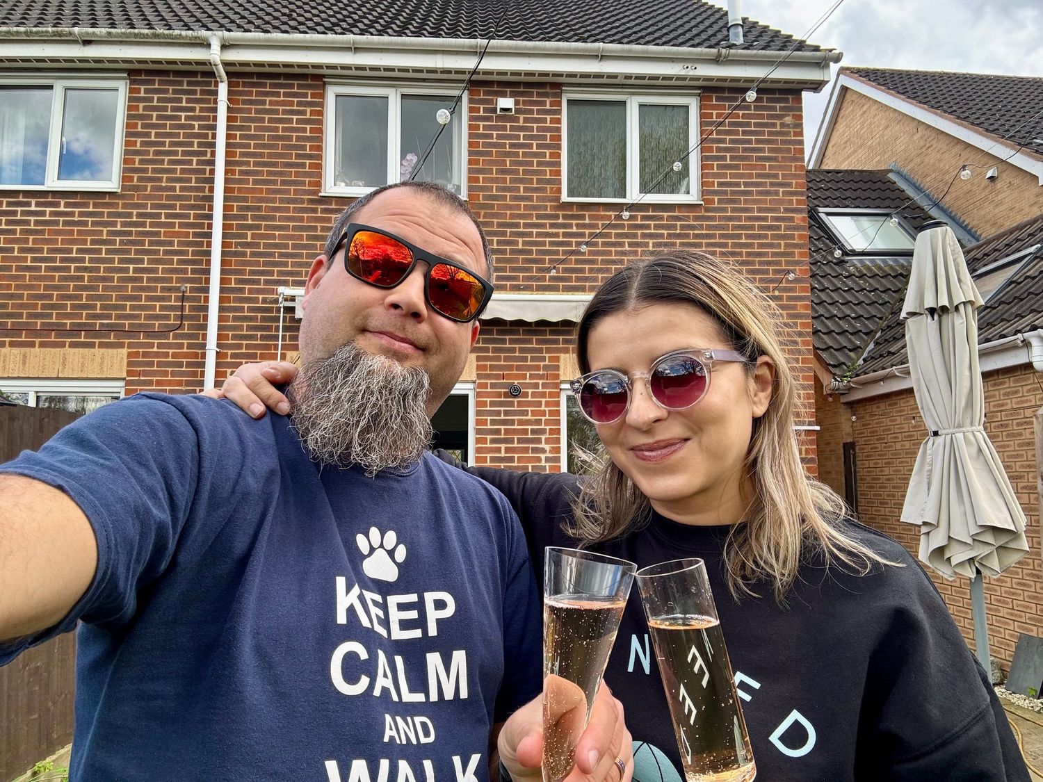 A casual outdoor selfie showing two people wearing sunglasses and holding wine glasses in front of a brick house.