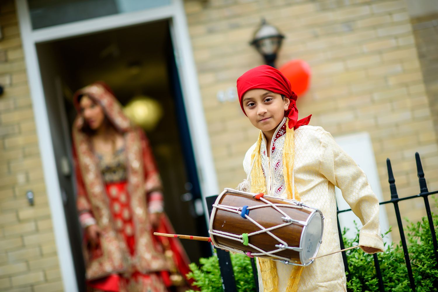 A young drummer in traditional attire performs with a dhol drum outside a brick building.