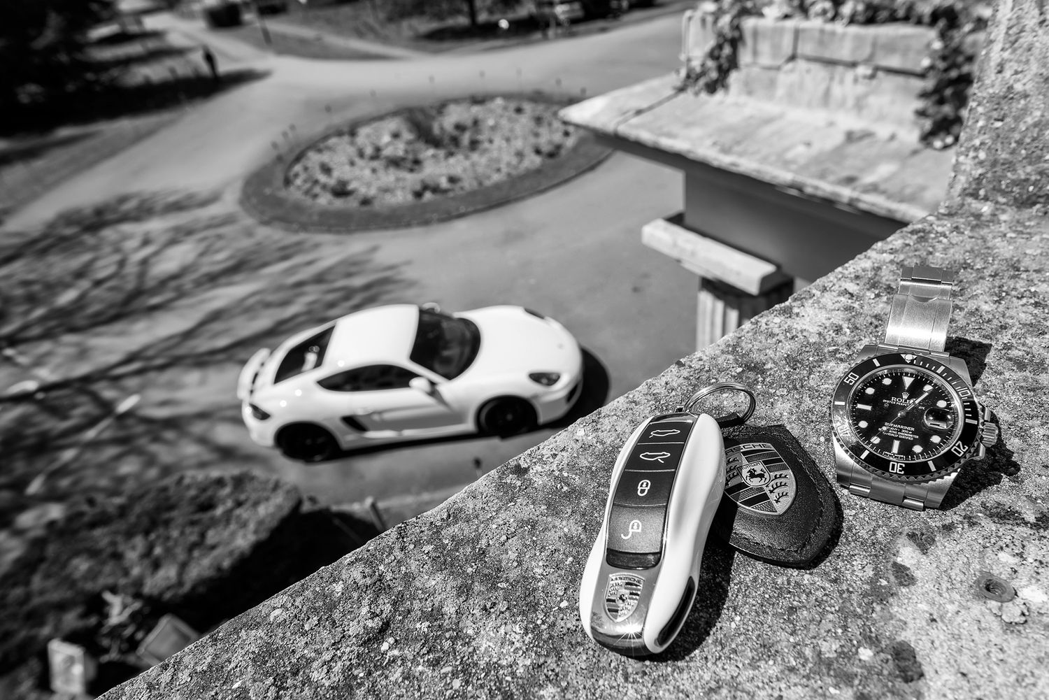 High-end luxury watch and car keys arranged next to a white sports car in monochromatic style.