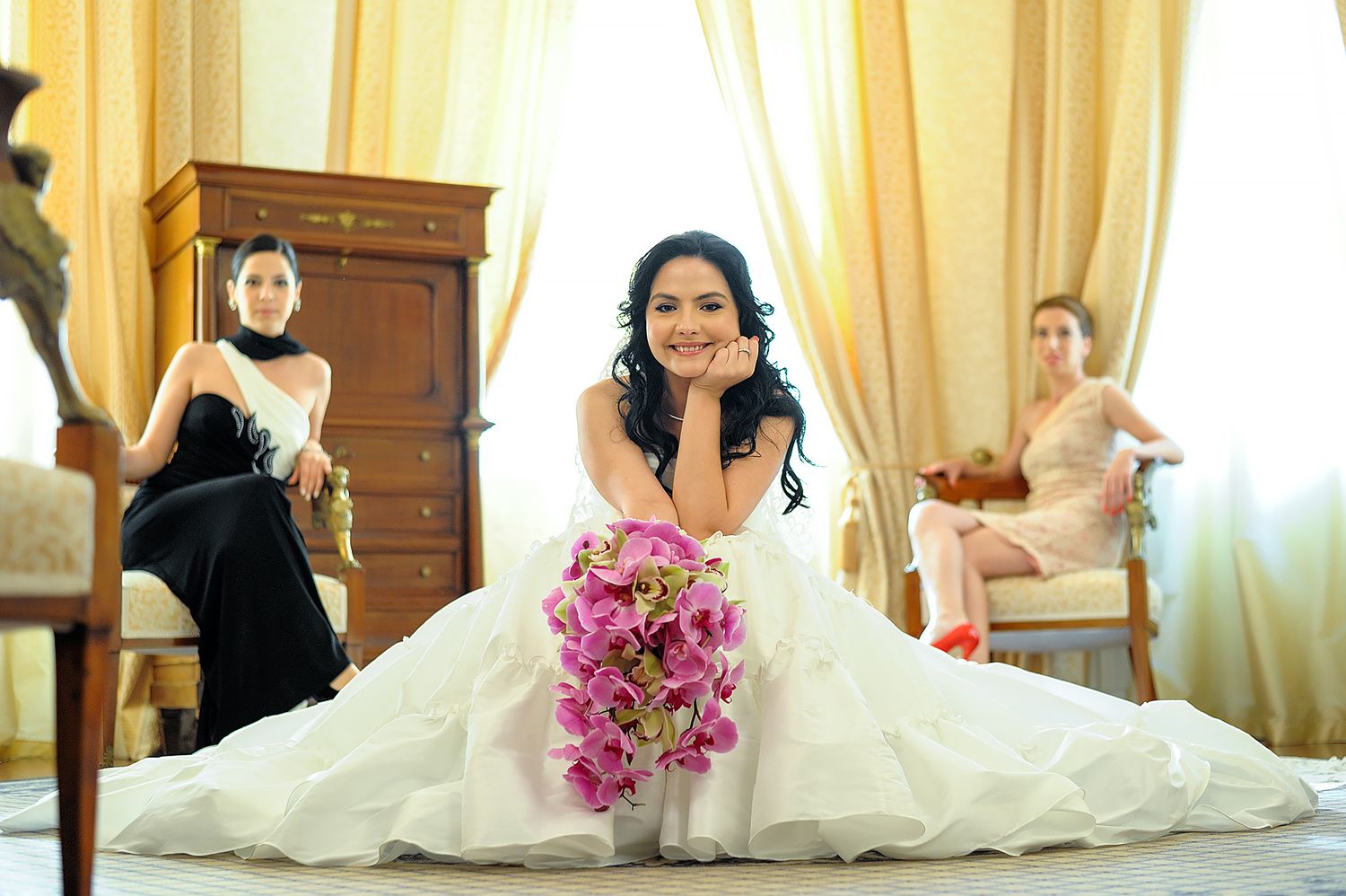 A bride in white dress poses with pink orchid bouquet in an elegant room.