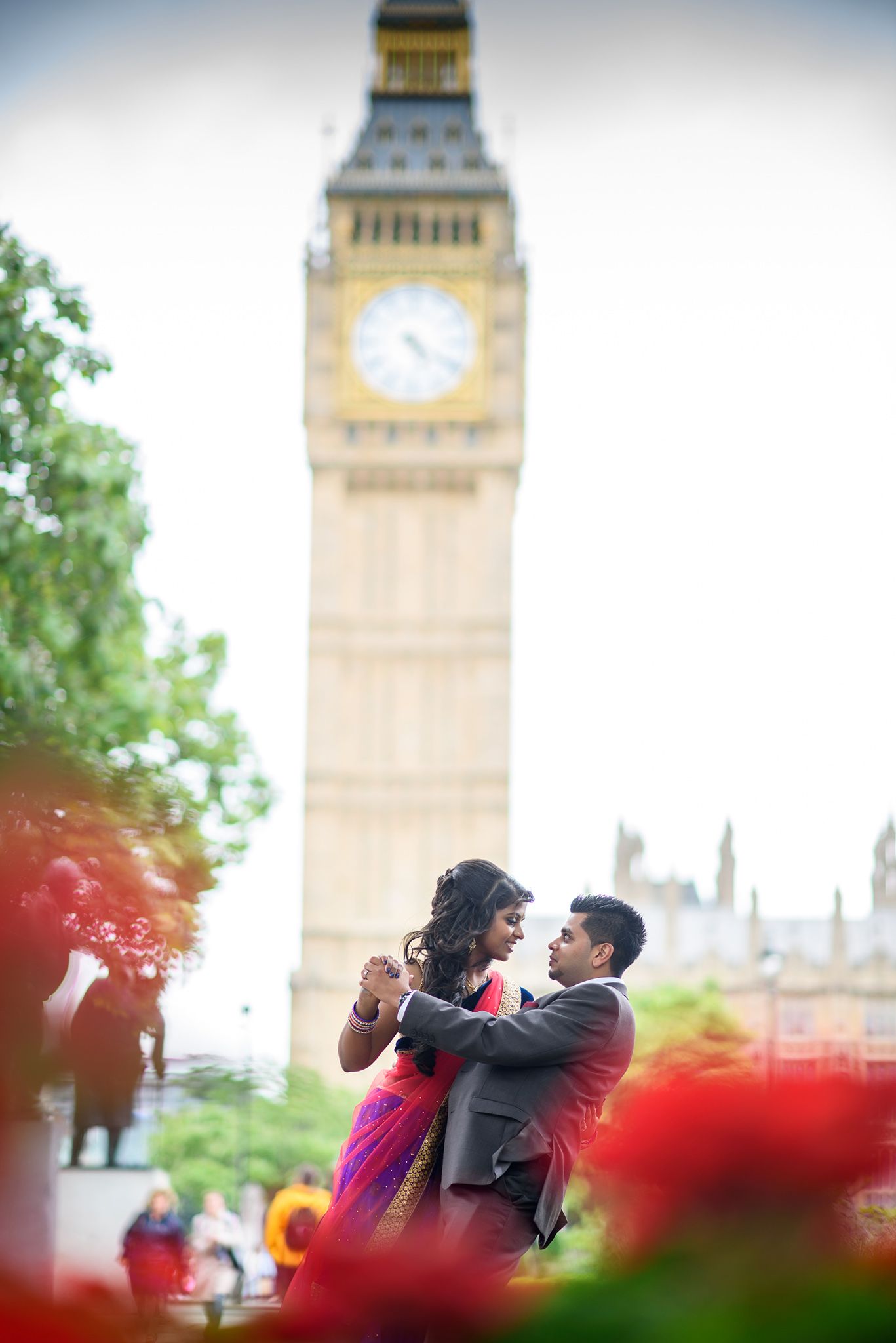 A romantic couple poses in front of London's Big Ben tower with red flowers in the foreground.