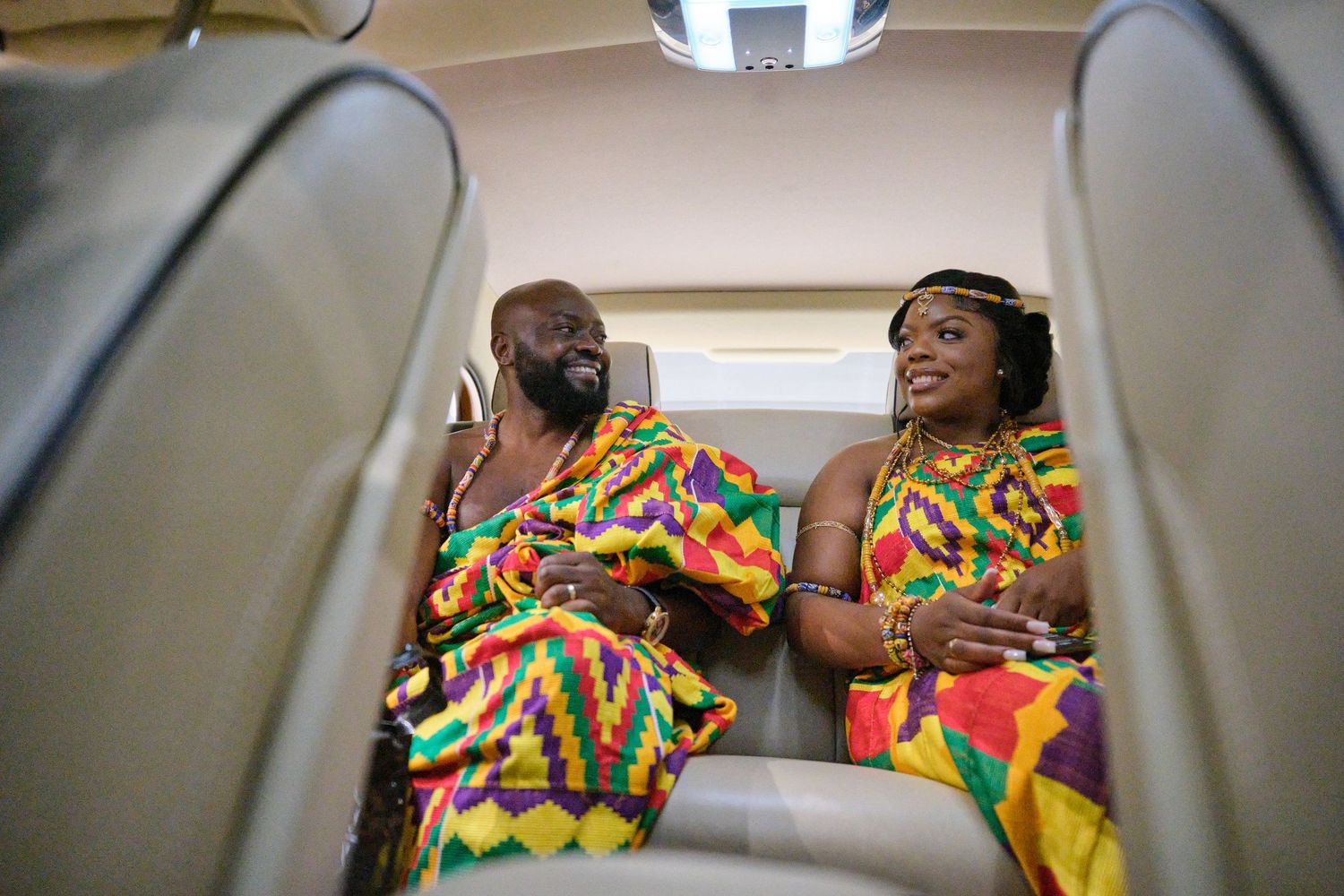 Two people wearing colorful African print outfits dance and celebrate in the back seat of a car.