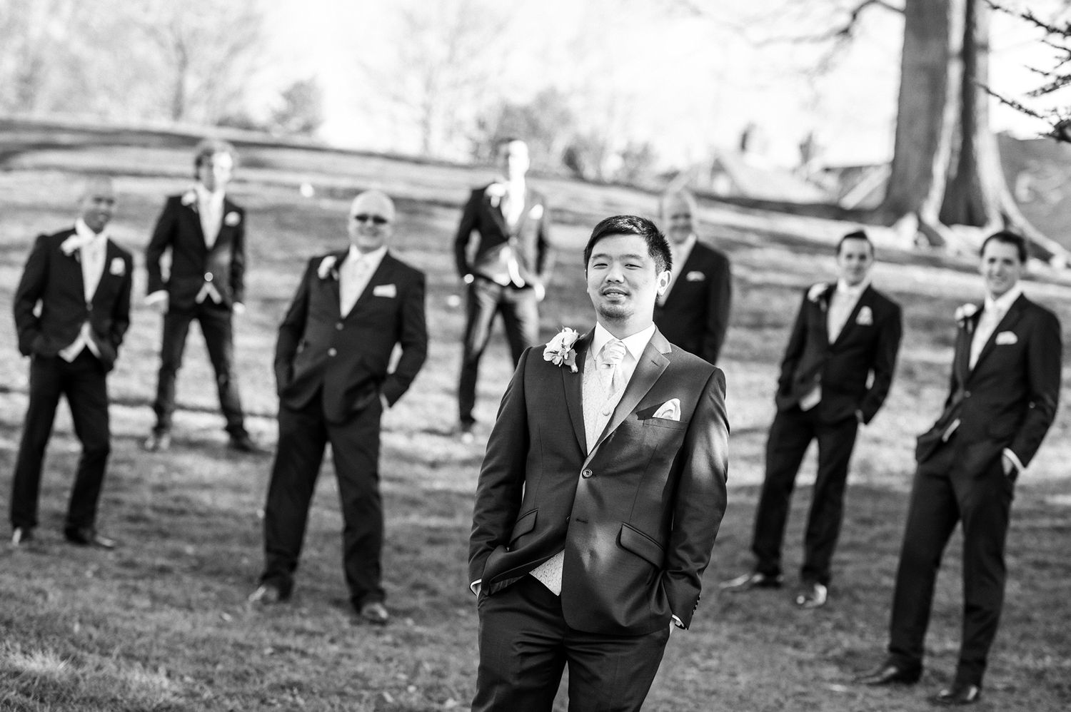 Groom stands confidently as groomsmen pose in background during outdoor wedding photoshoot.