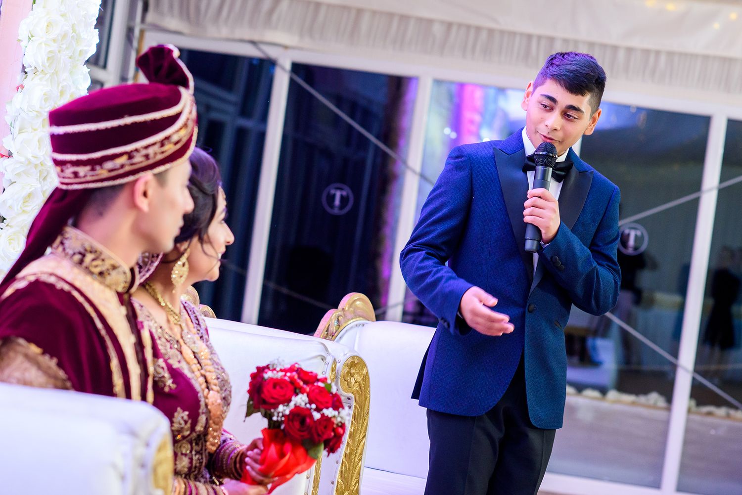 A guest gives a speech at a traditional Indian wedding ceremony with vibrant red decor.