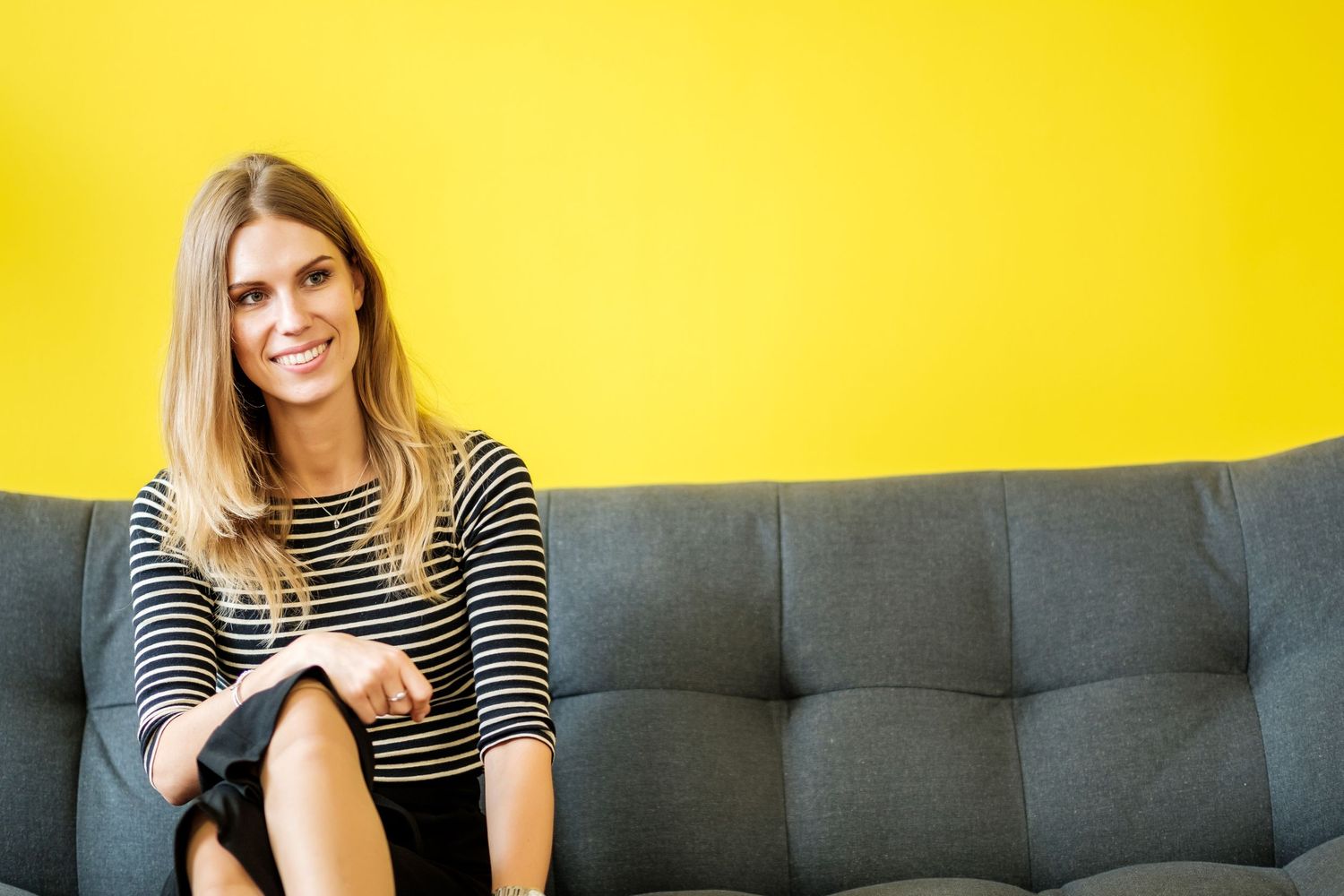 Woman in striped shirt sits casually on grey couch against vibrant yellow wall.