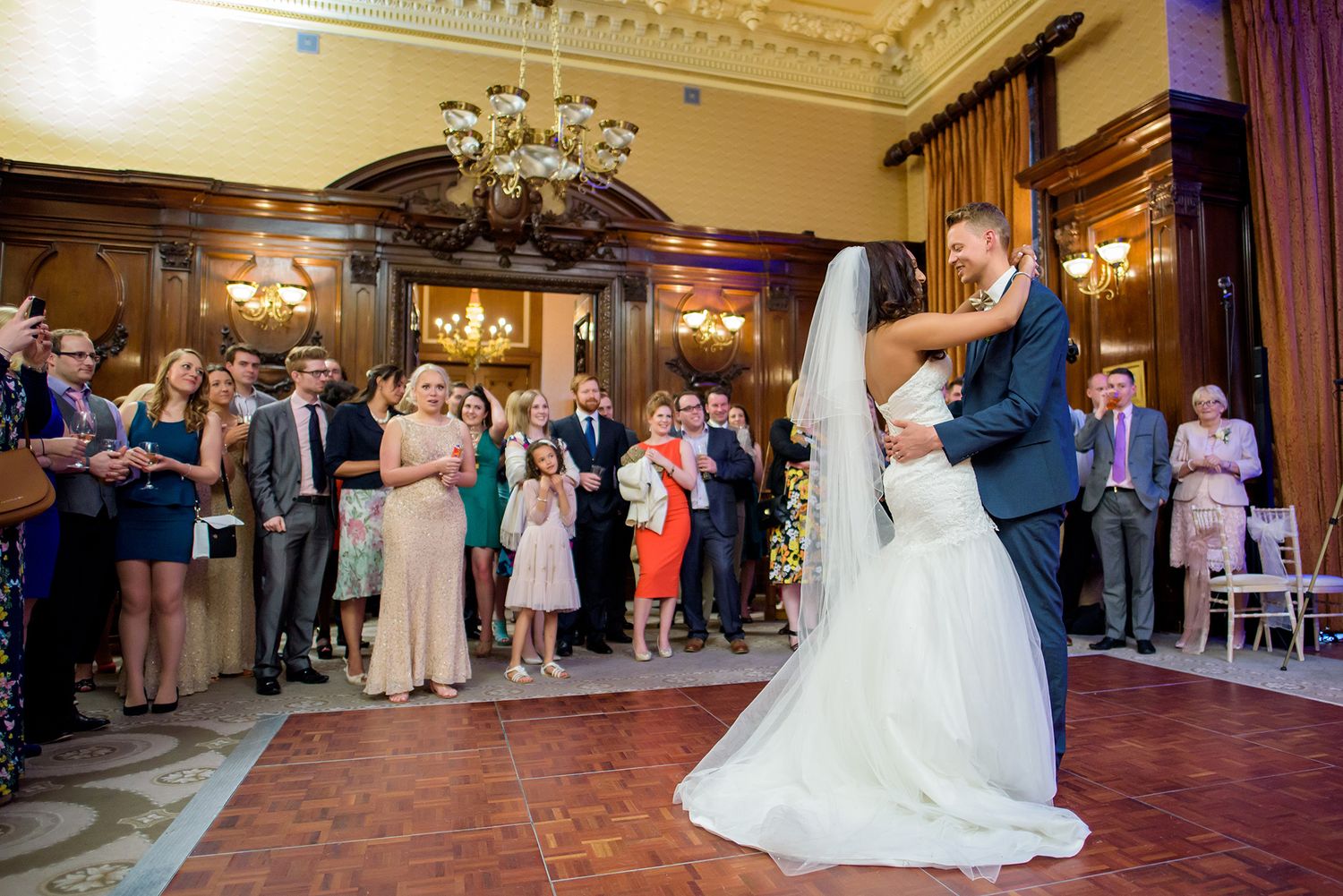 Couple sharing their first dance surrounded by guests in an ornate ballroom with chandelier.