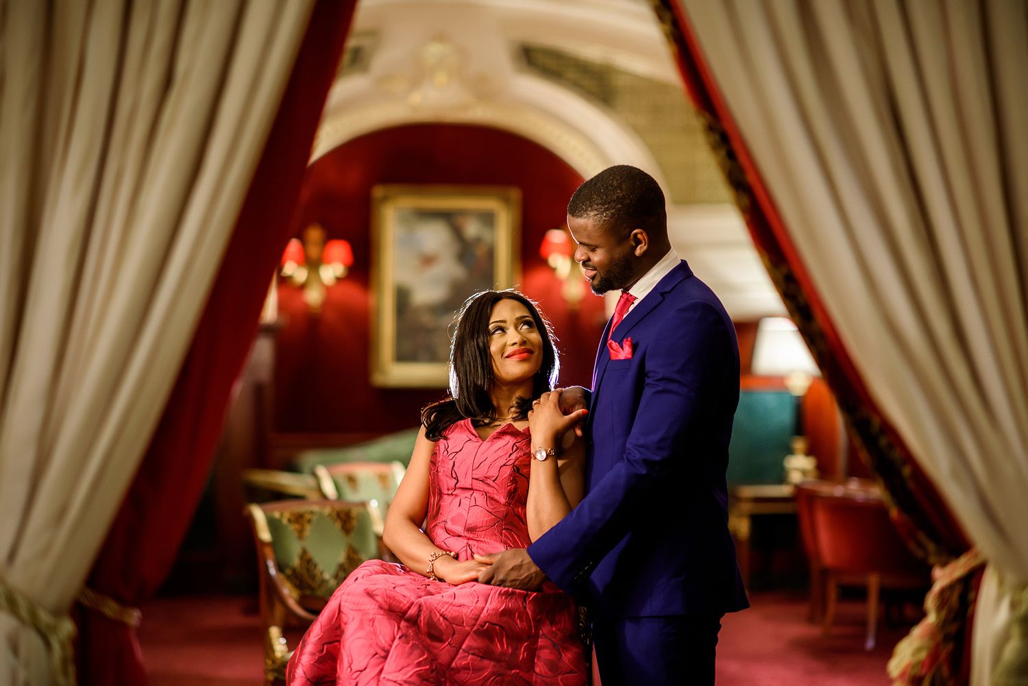 A couple shares an intimate moment in an elegantly decorated room with red curtains and warm lighting.
