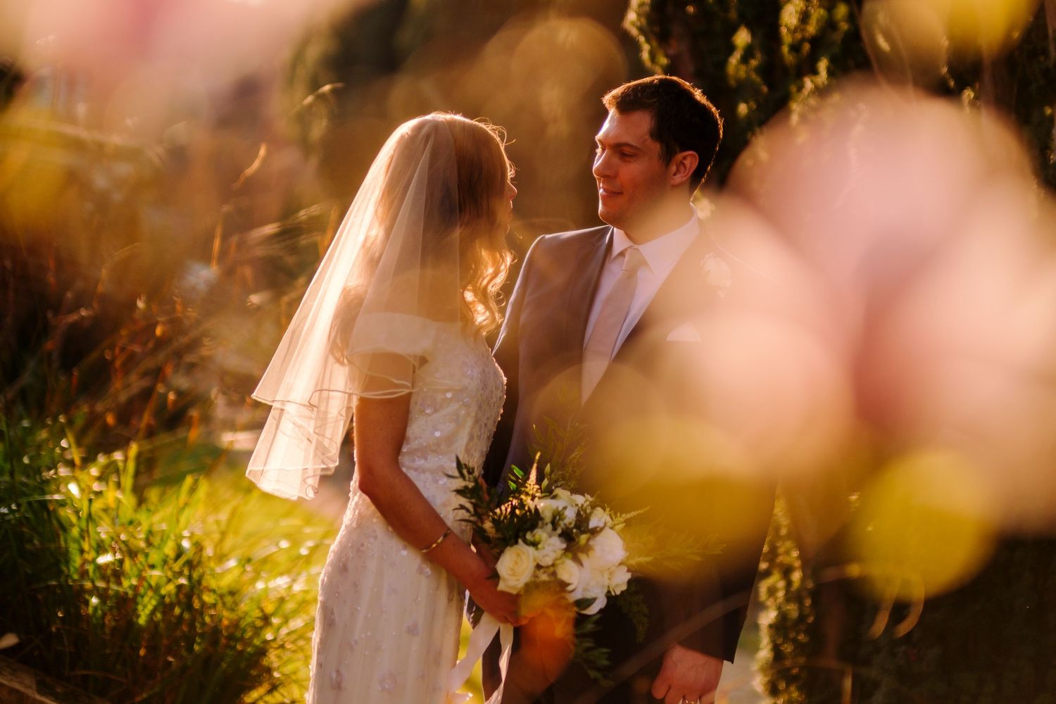 Romantic sunset wedding portrait through soft bokeh flowers.