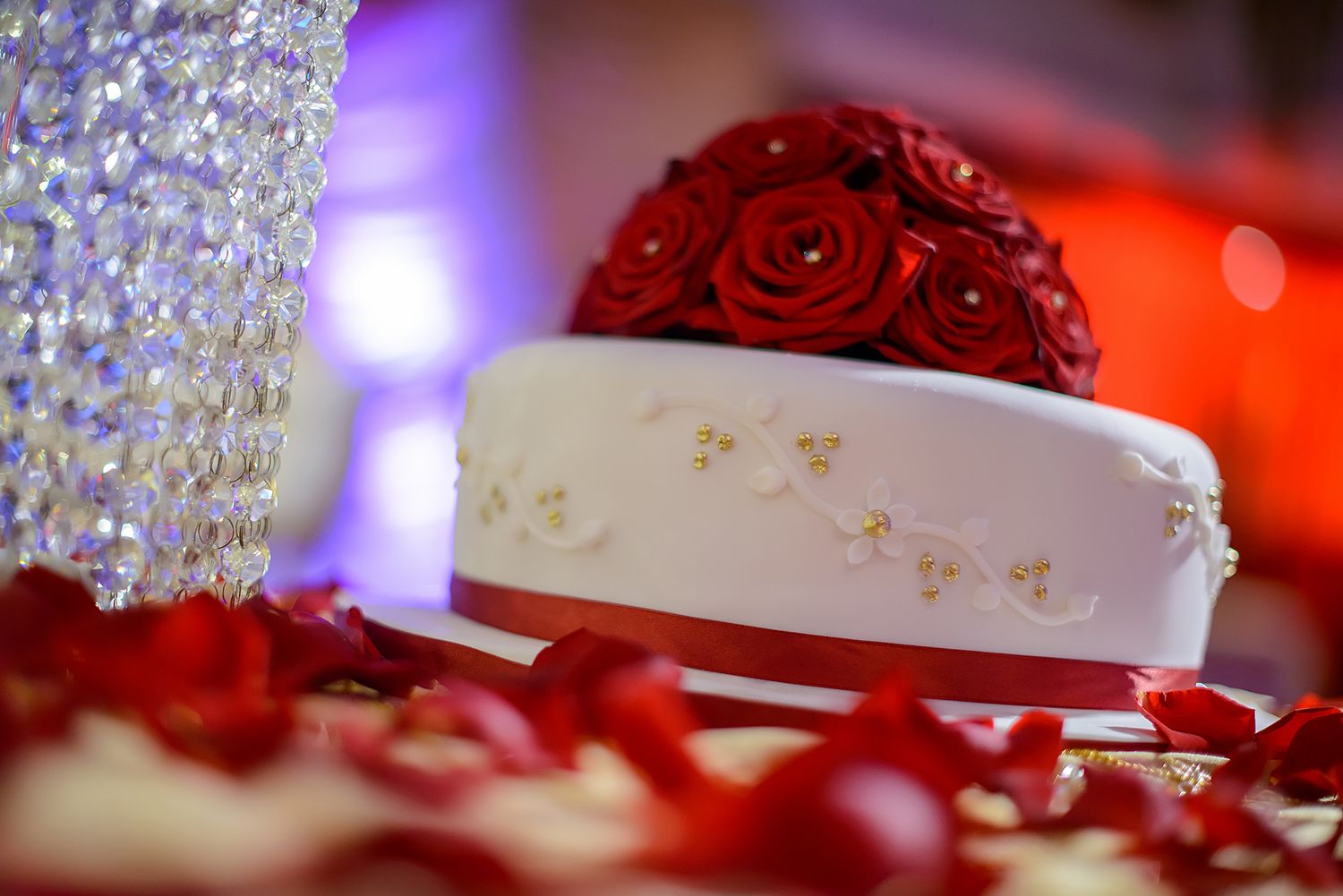 Elegant wedding cake decorated with red roses and crystal accents under purple lighting.