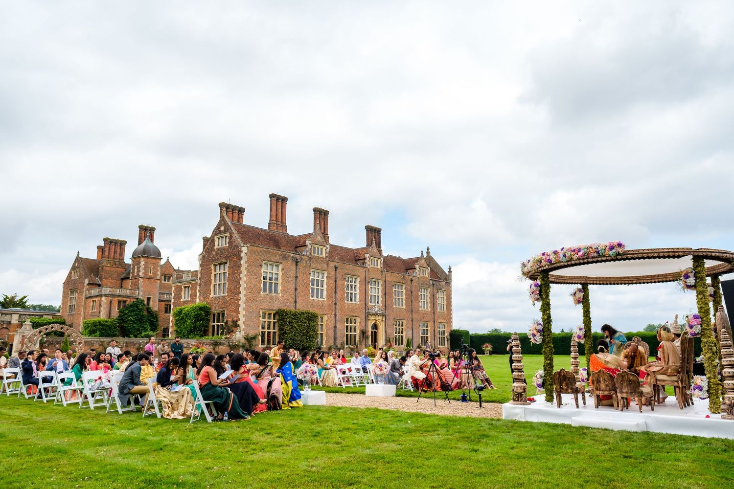 Panoramic view of wedding guests seated outdoors at a historic brick manor house under cloudy skies.