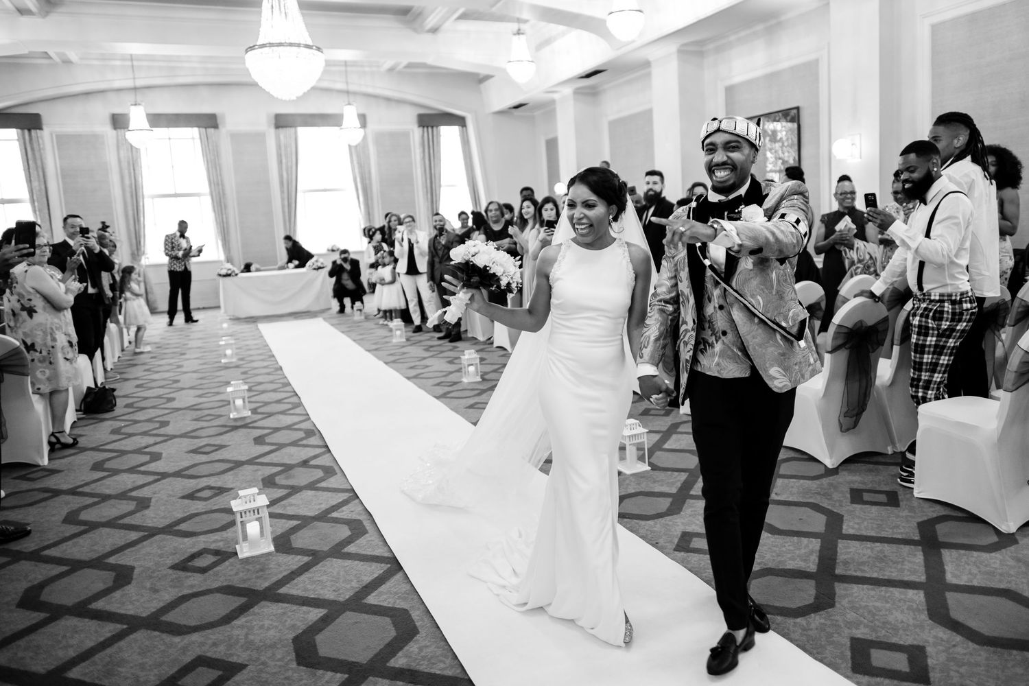Wedding reception guests dancing and celebrating in an elegant white ballroom with patterned carpet.