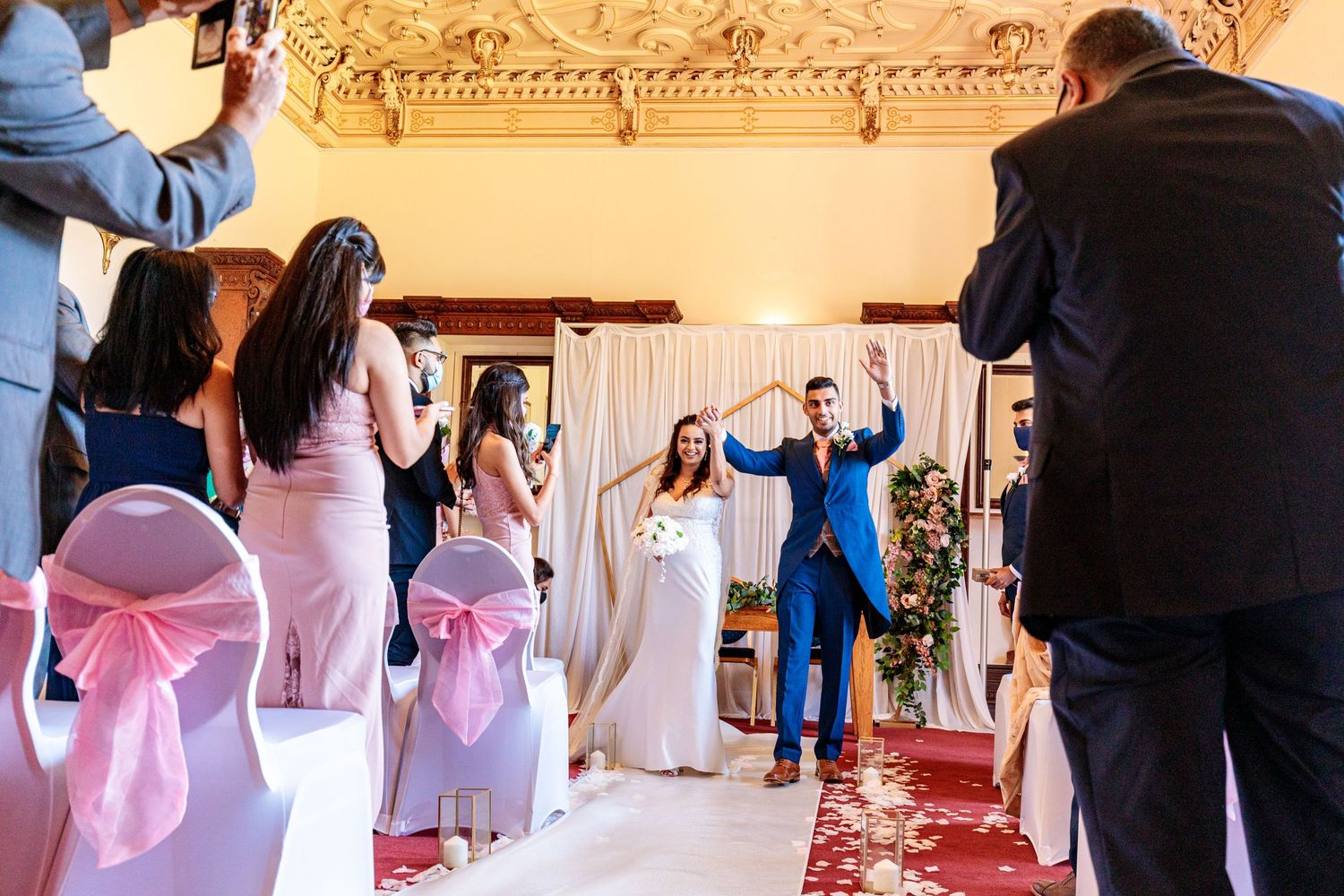 Wedding ceremony setup in an elegant ballroom with pink chair sashes and ornate architectural details.