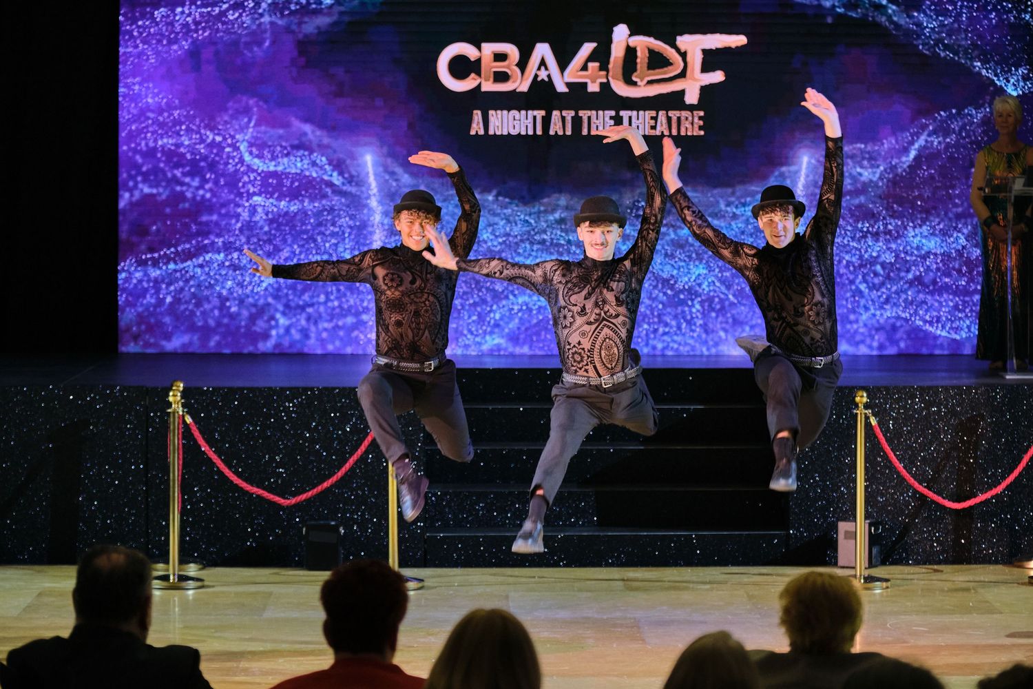 Theater stage performance with dramatic purple lighting and performers in dark attire.