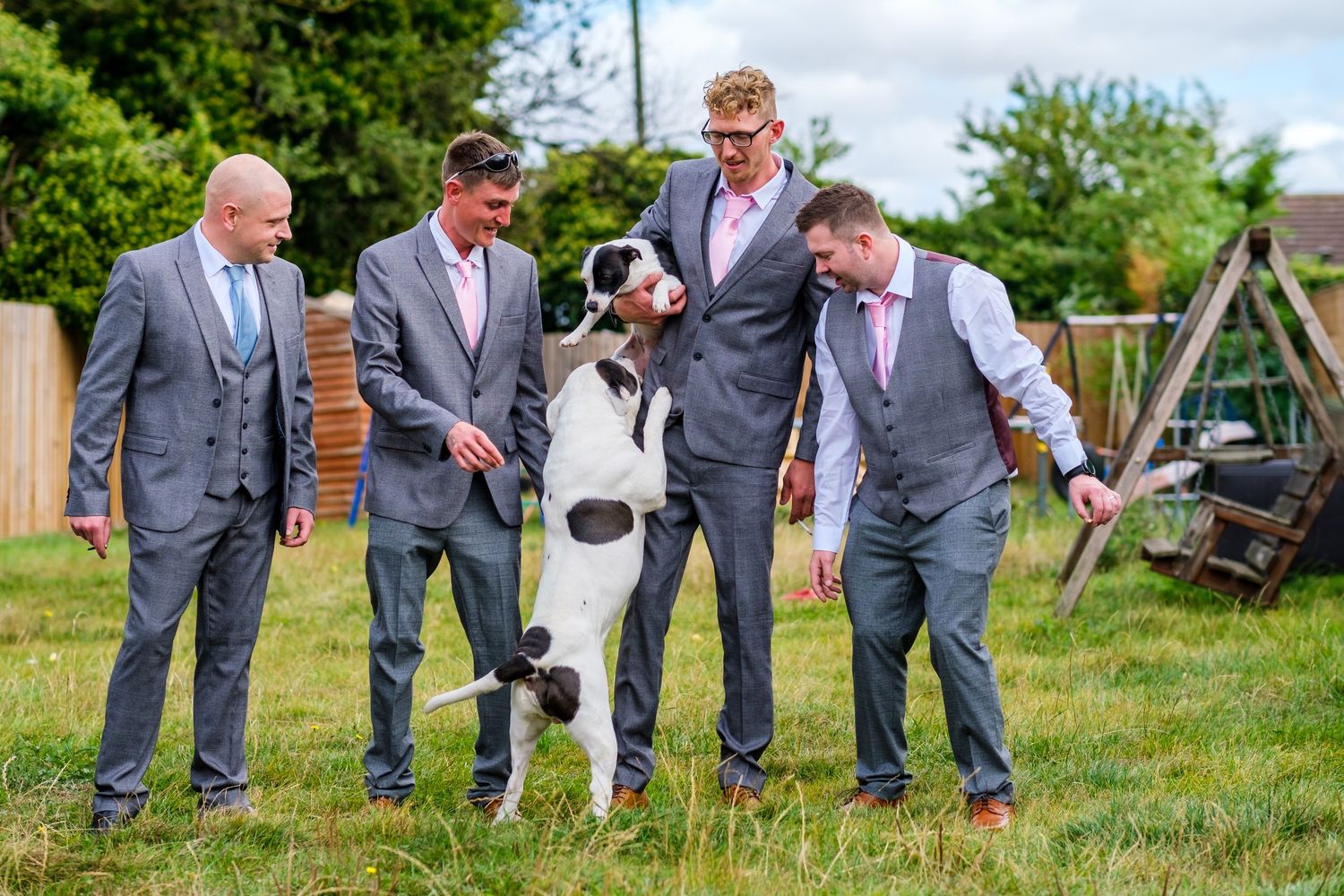 Group of men in grey suits playing with a black and white dog on grass.