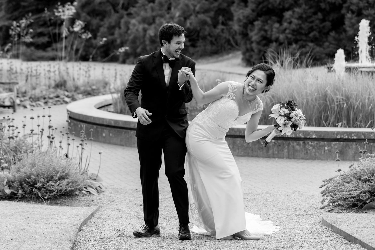 A joyful wedding couple shares a candid moment together in a garden setting, captured in black and white.