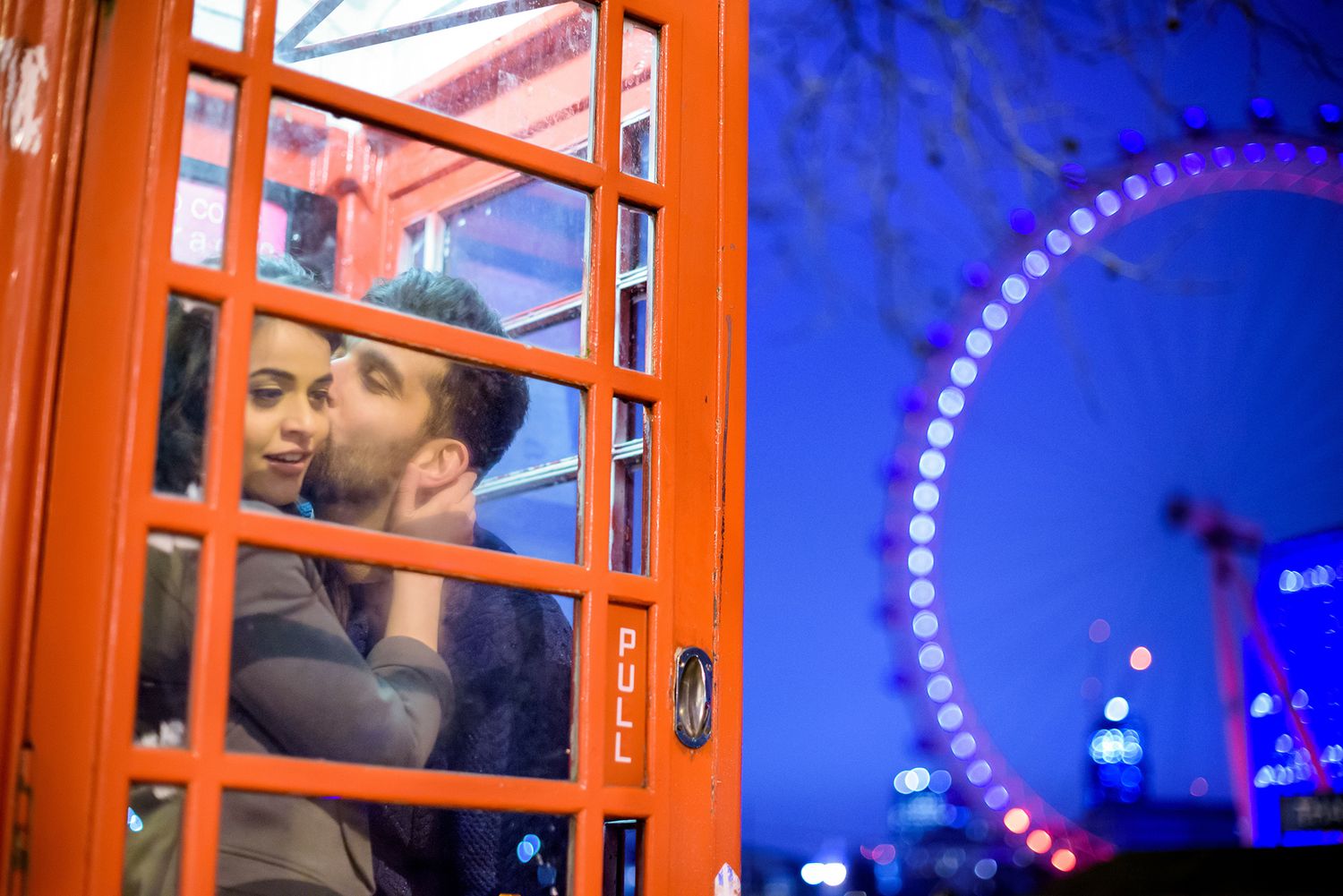 Red phone booth frames romantic moment against London Eye at twilight.