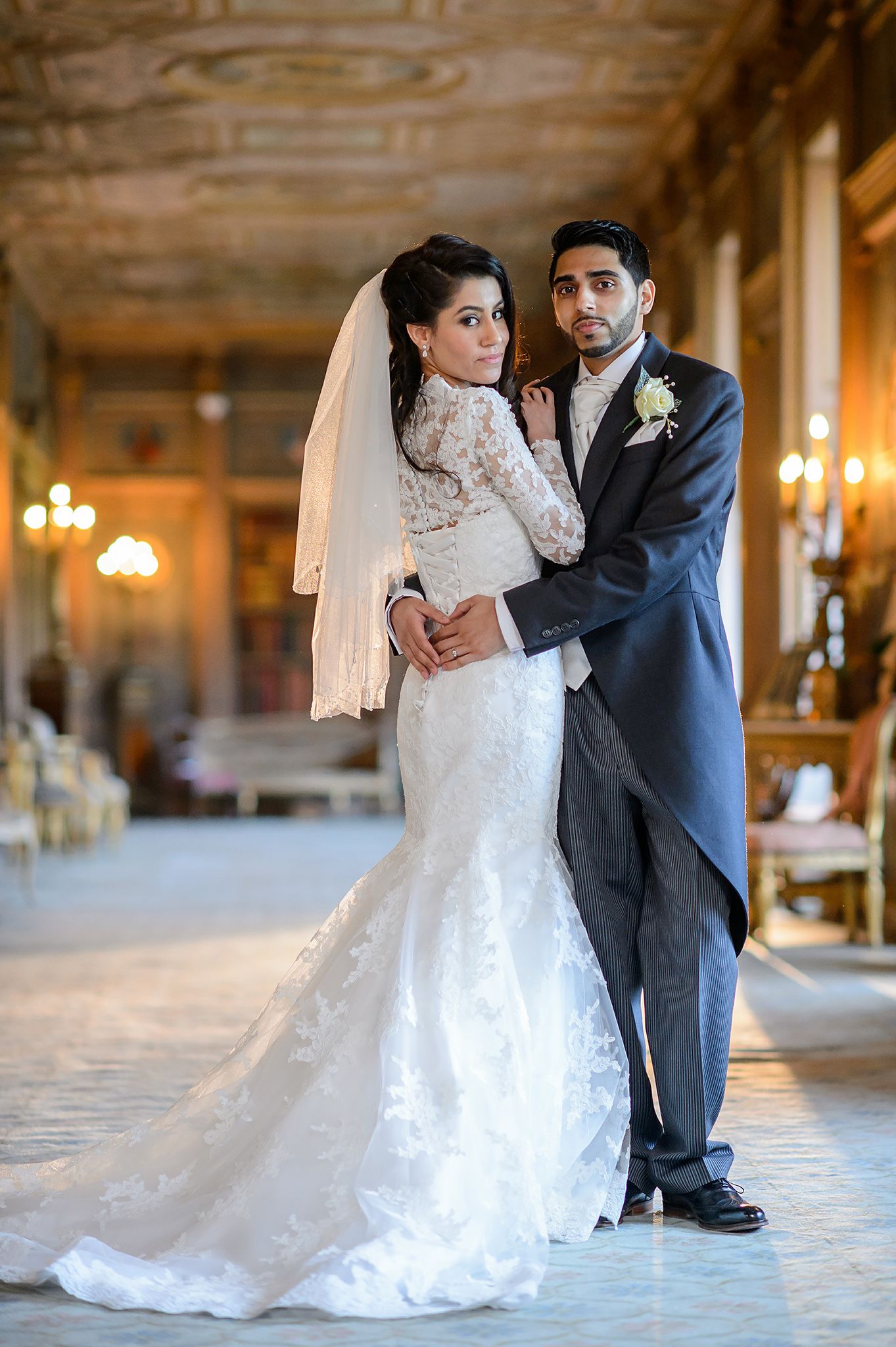 Wedding couple in elegant attire share romantic pose in luxurious hall with chandeliers.