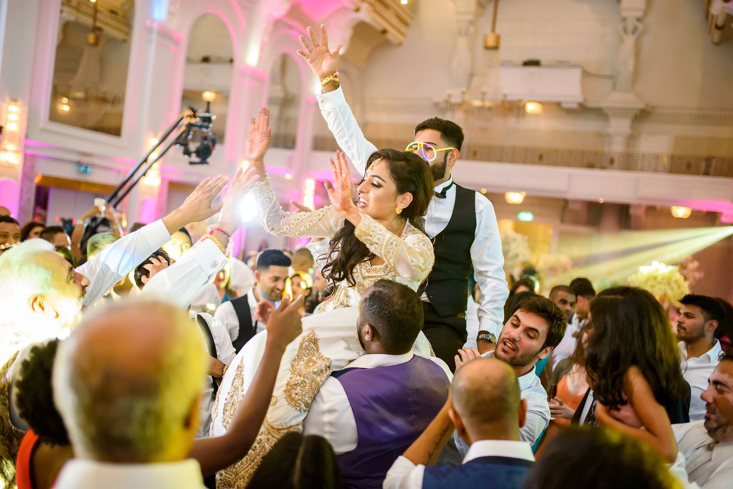 Large crowd of wedding guests dancing and celebrating in an ornate ballroom venue.