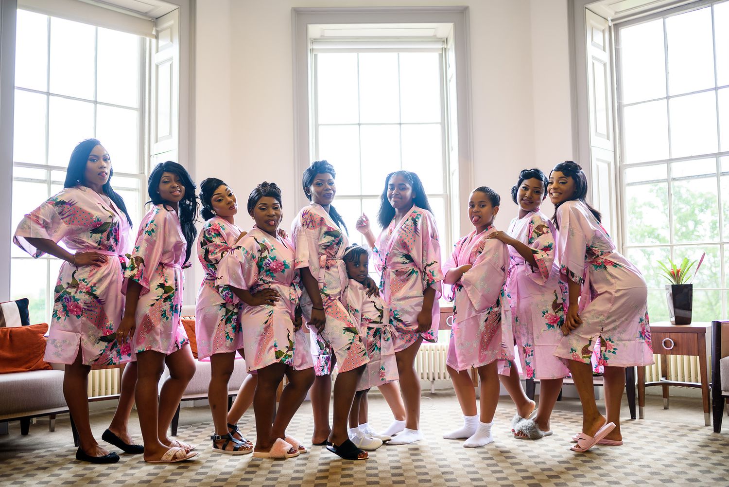 Line of bridesmaids in pink dresses pose together in an elegant room with large windows.
