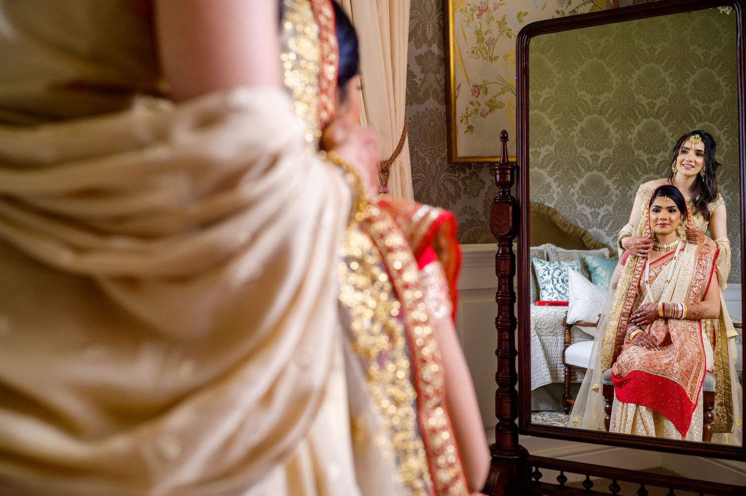 Traditional Indian bridal preparation scene reflected in a mirror with ornate wallpaper backdrop.