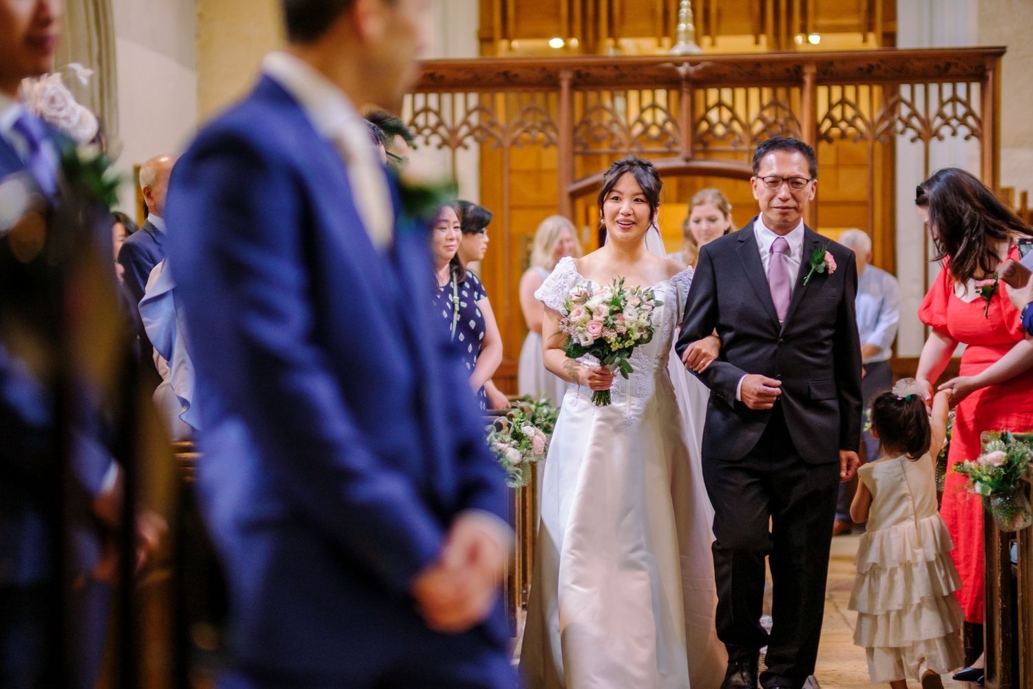 Wedding ceremony guests stand in an ornate church with beautiful stained glass windows and wooden pews.