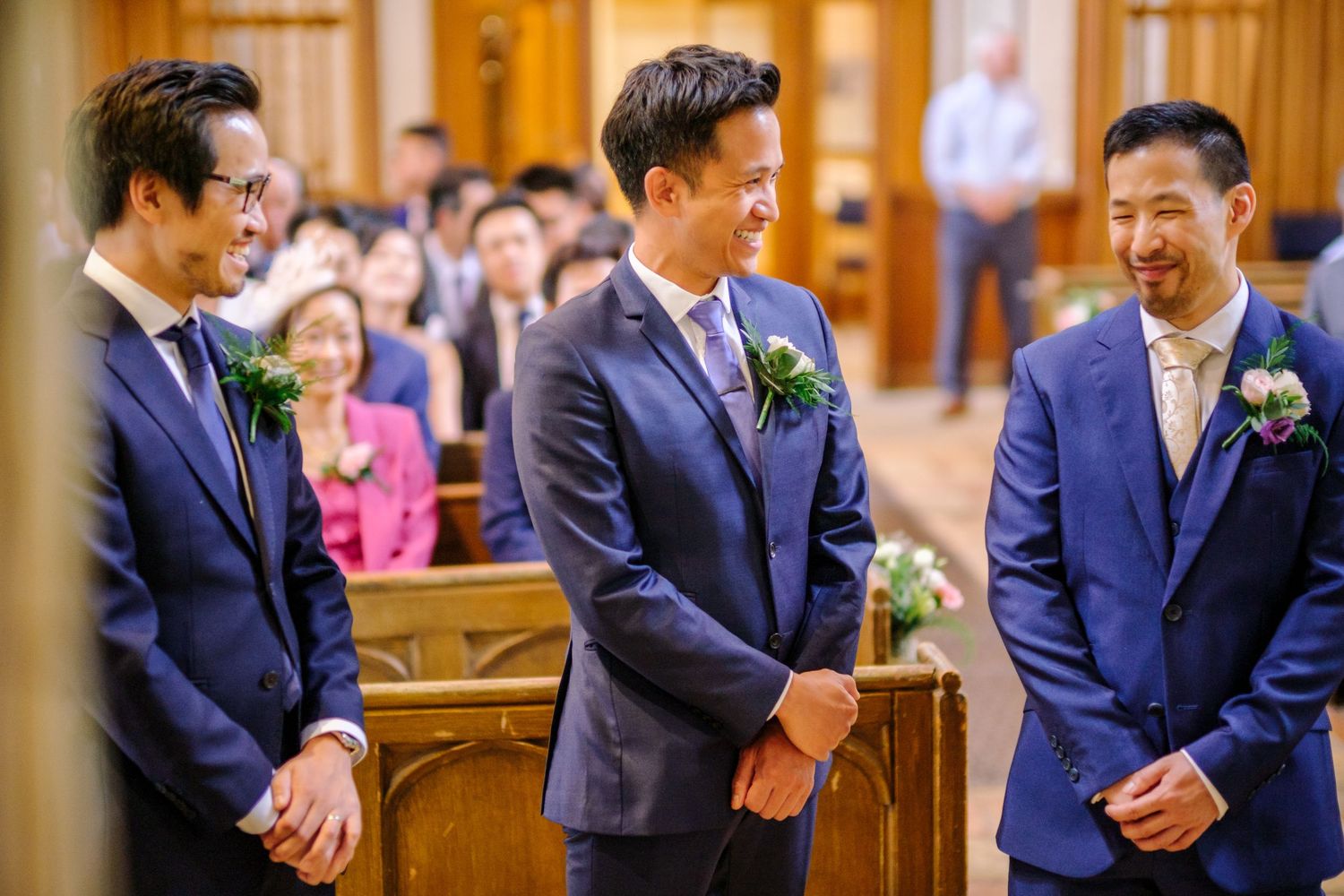 Groomsmen in navy blue suits share a lighthearted moment while standing in a church during a wedding ceremony.