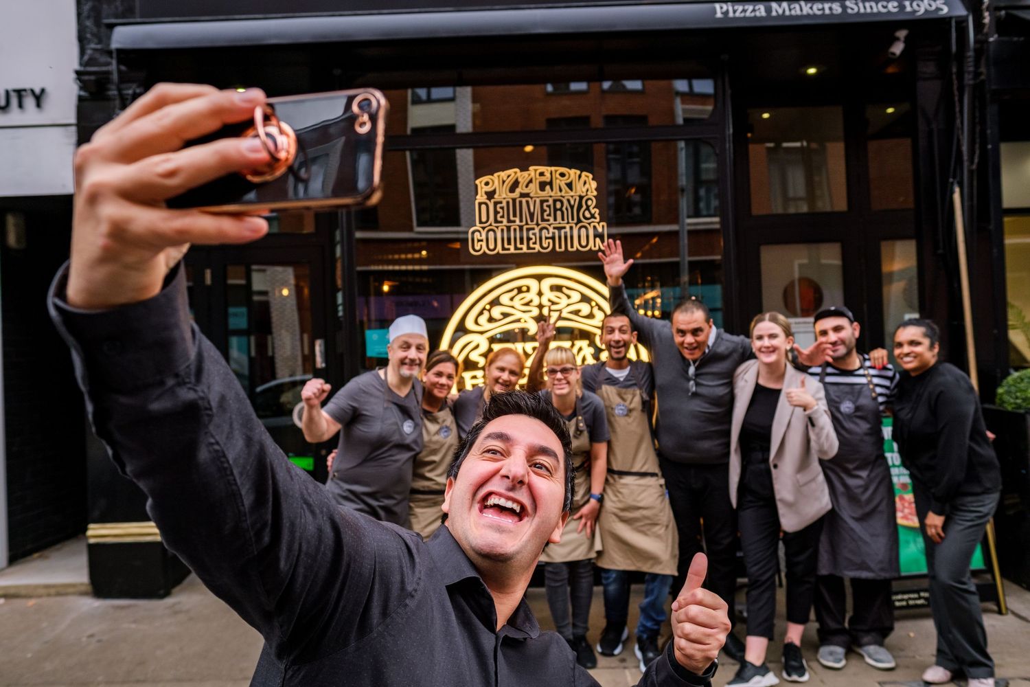 Group of excited customers taking selfies outside a restaurant with illuminated signage.