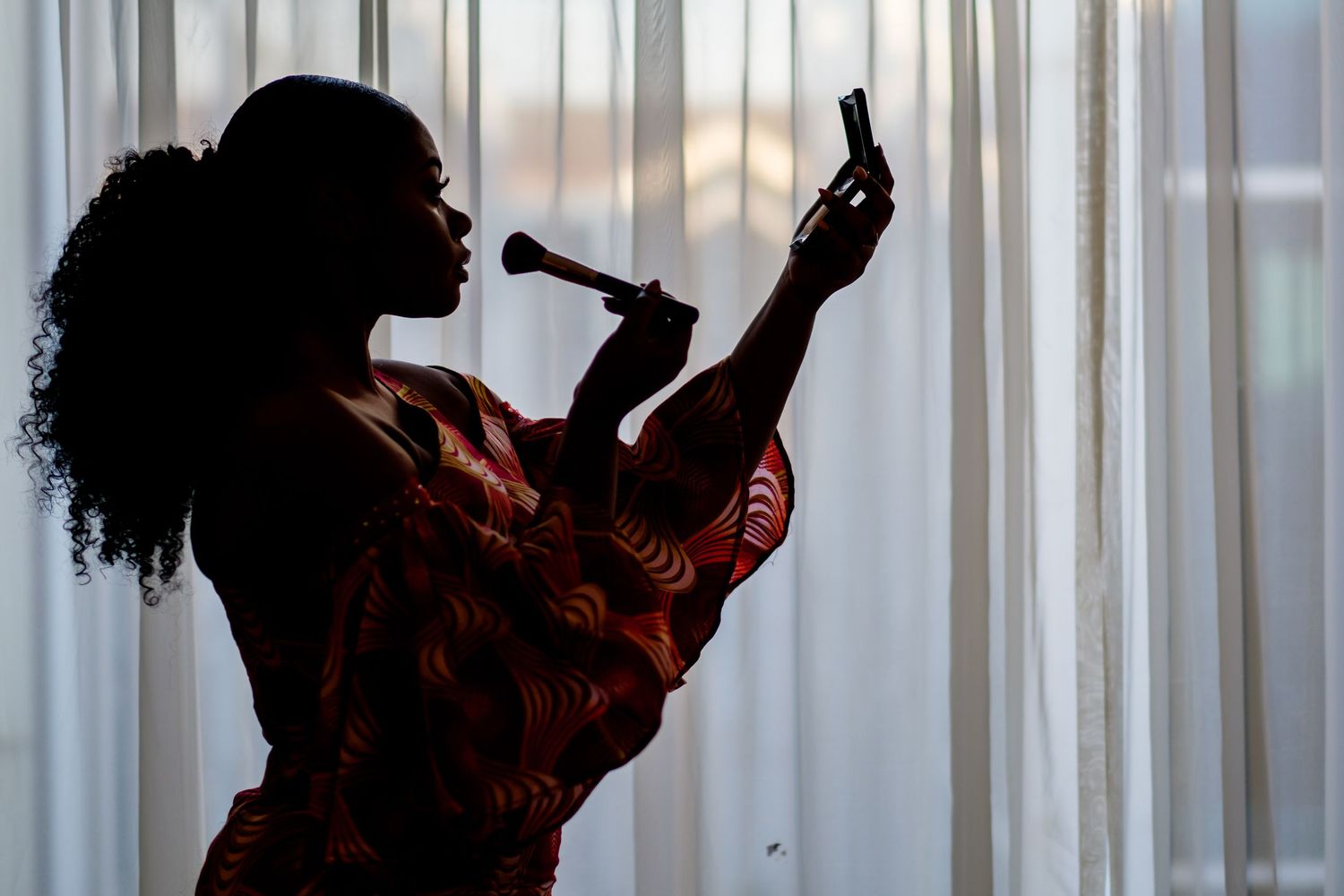 Silhouette against window blinds holding an object up to their face.