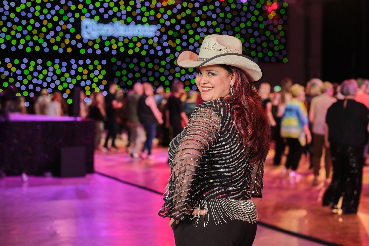 Country western dancer in a cowboy hat and fringed jacket spins on a purple lit dance floor.
