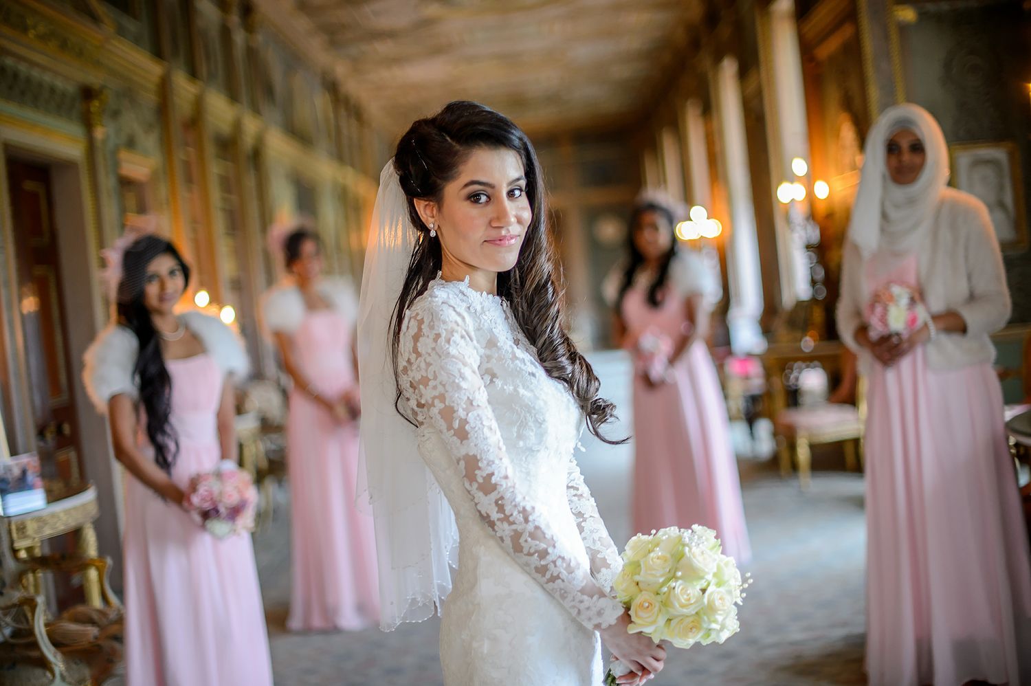 Bride in lace dress holds white bouquet while bridesmaids in pink dresses stand in background.