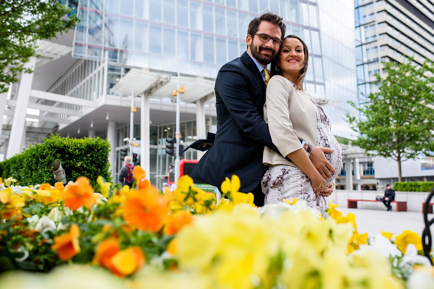 Cheerful couple embracing outdoors with yellow flowers and modern architecture in background.