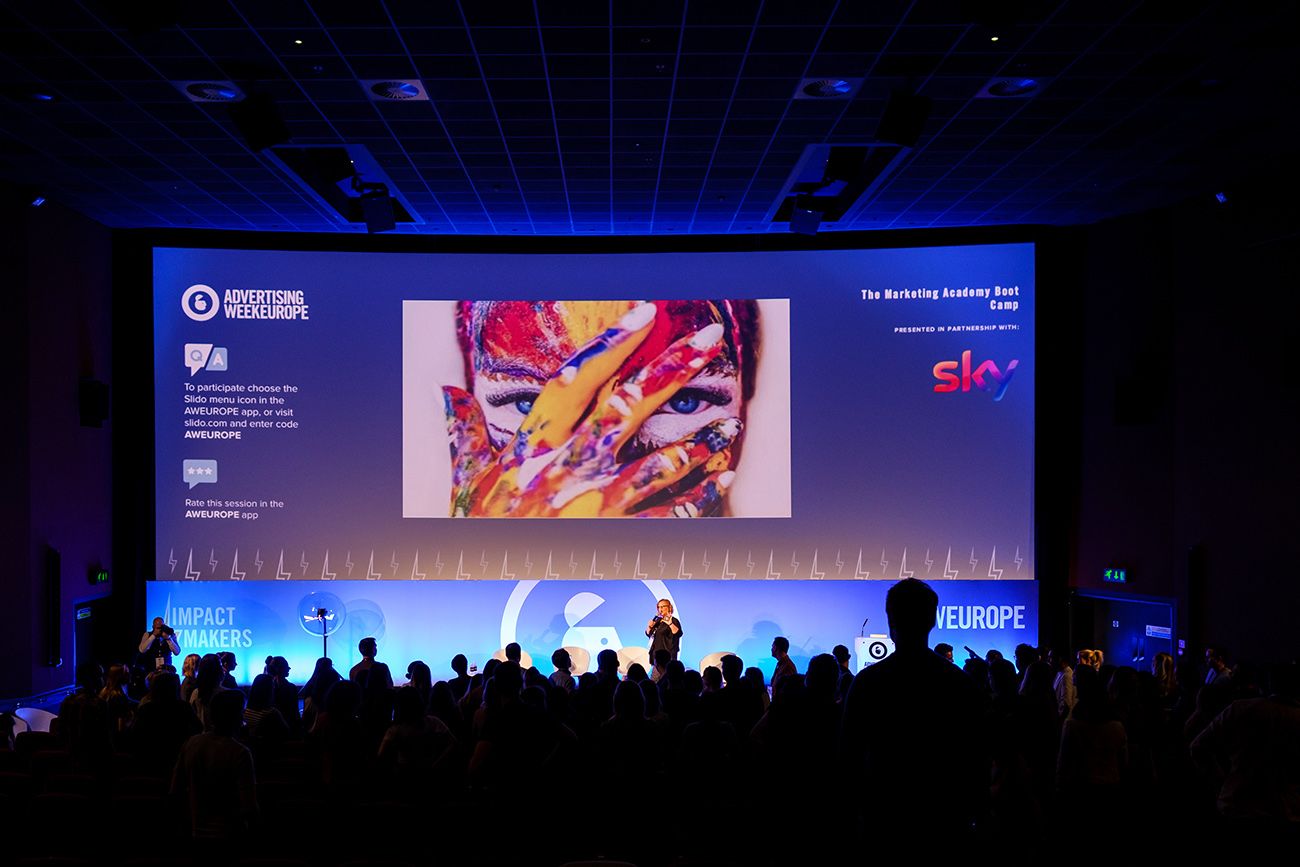 Wide shot of a dark conference room with blue lighting and audience silhouettes against large presentation screens.