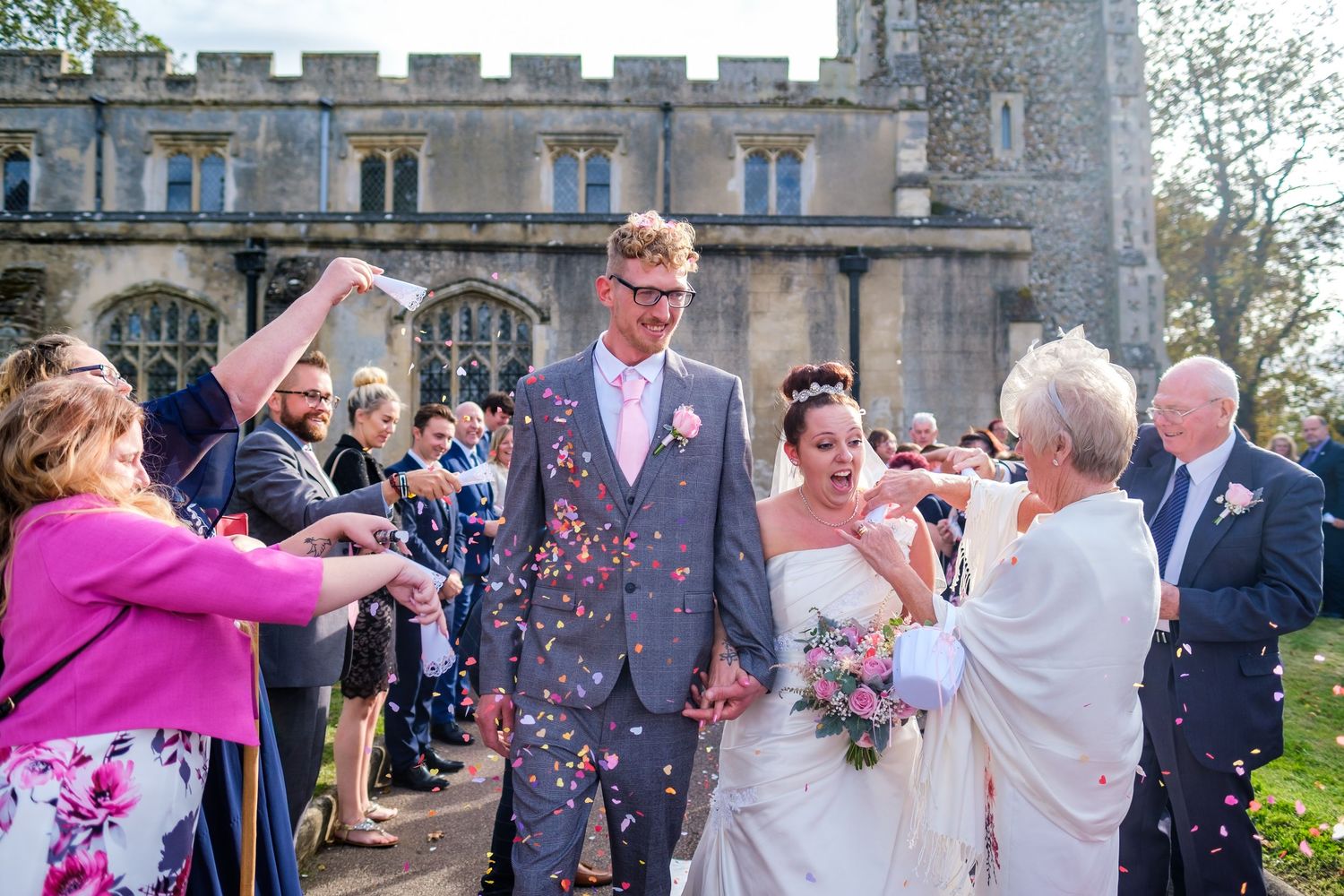 Guests throw confetti at a joyful wedding celebration outside a stone church.