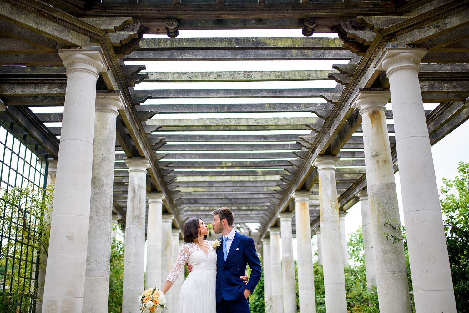 Wedding couple poses under elegant white columned pergola surrounded by lush greenery.