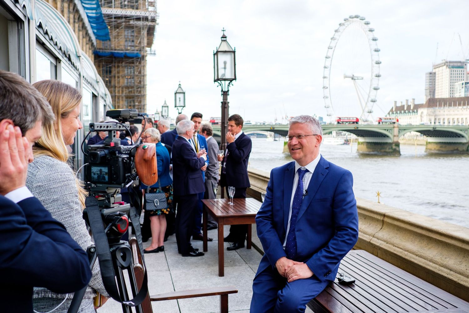 People gathered on a riverside terrace with the London Eye visible in the background across the Thames.