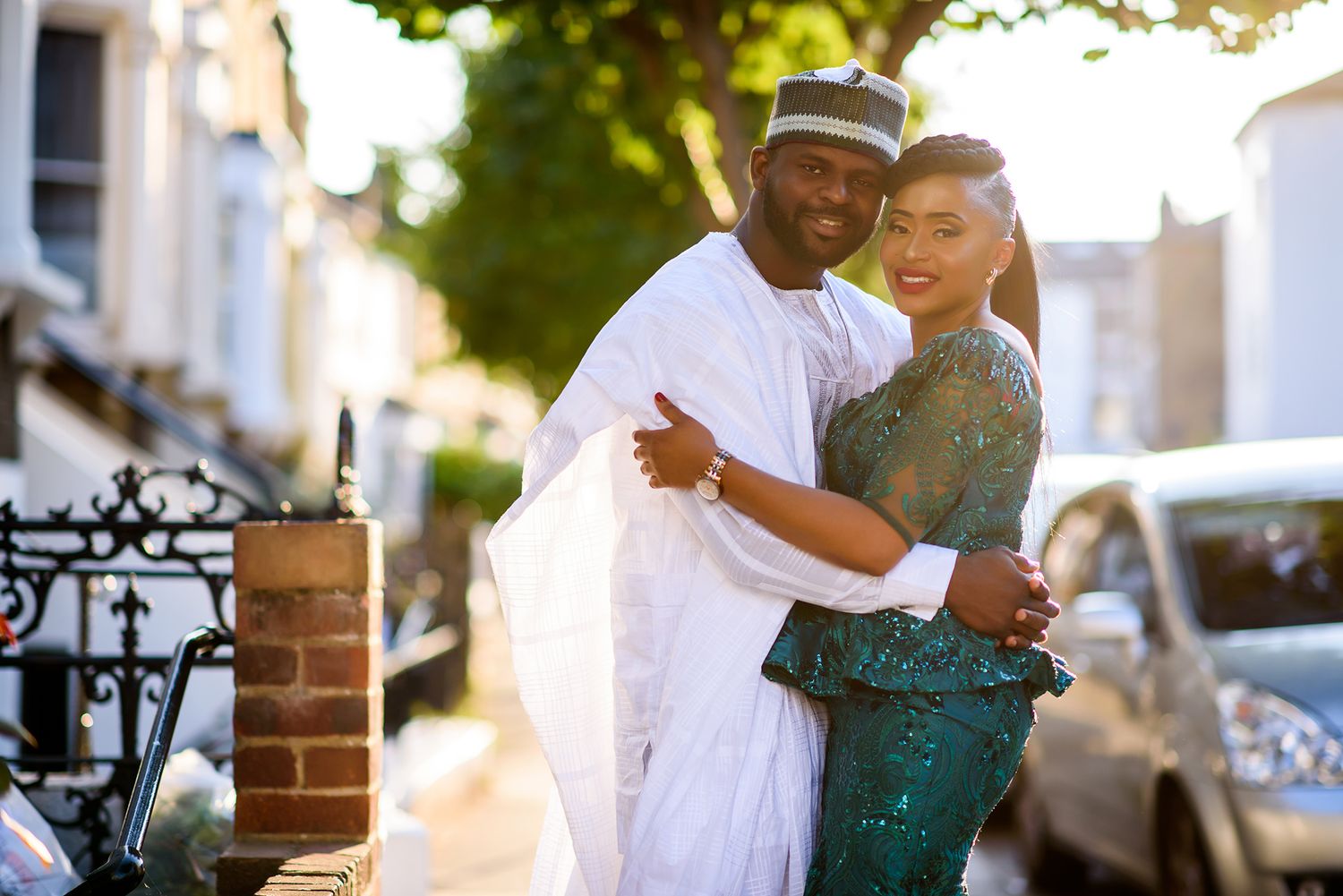 A couple in elegant attire share a romantic moment beneath lush green trees on a sunny afternoon.