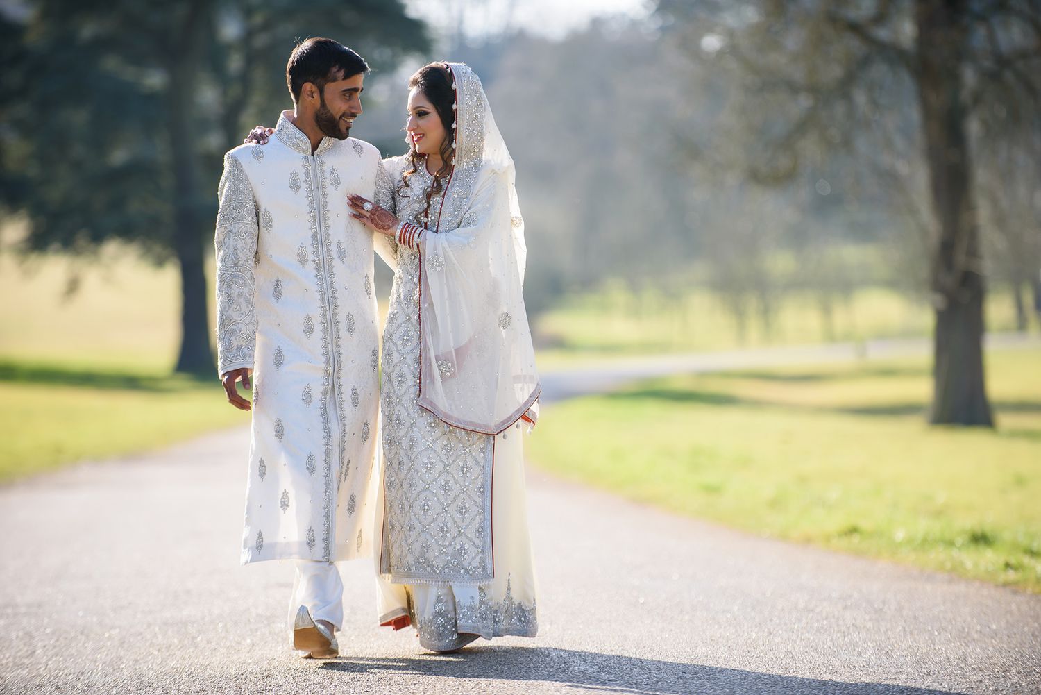 Couple in traditional white South Asian attire walking together on a sunlit path through a park.