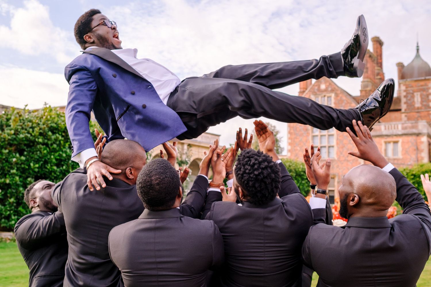 Groomsmen in dark suits pose playfully while lying on grass with church spires in background.