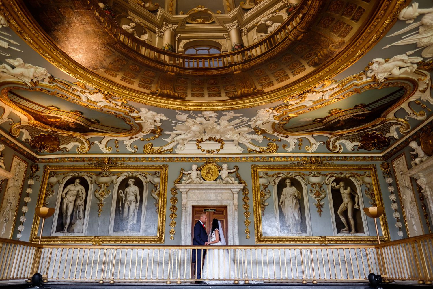 Wedding photo in an opulent room with gold-detailed ceiling and classical artwork.