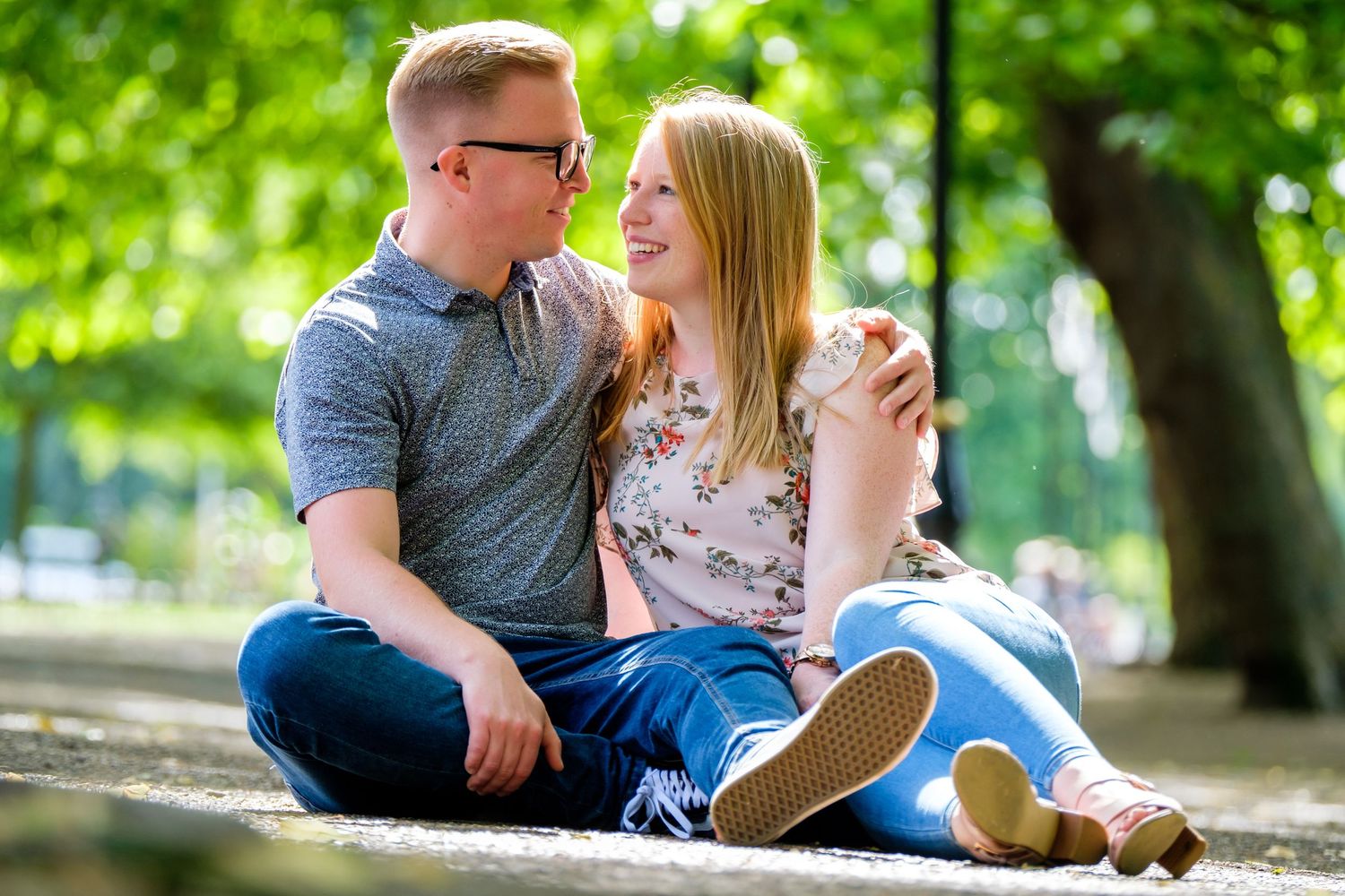 A romantic couple sits together on the ground in a sunny park setting.