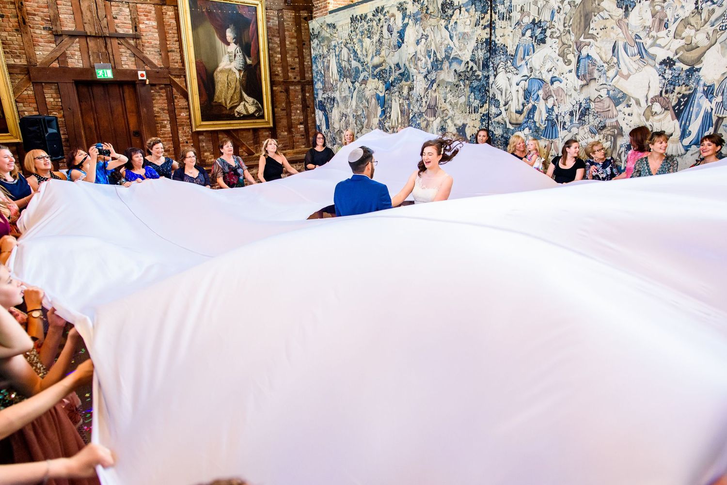 Wedding guests lifting a white parachute during an indoor reception celebration.