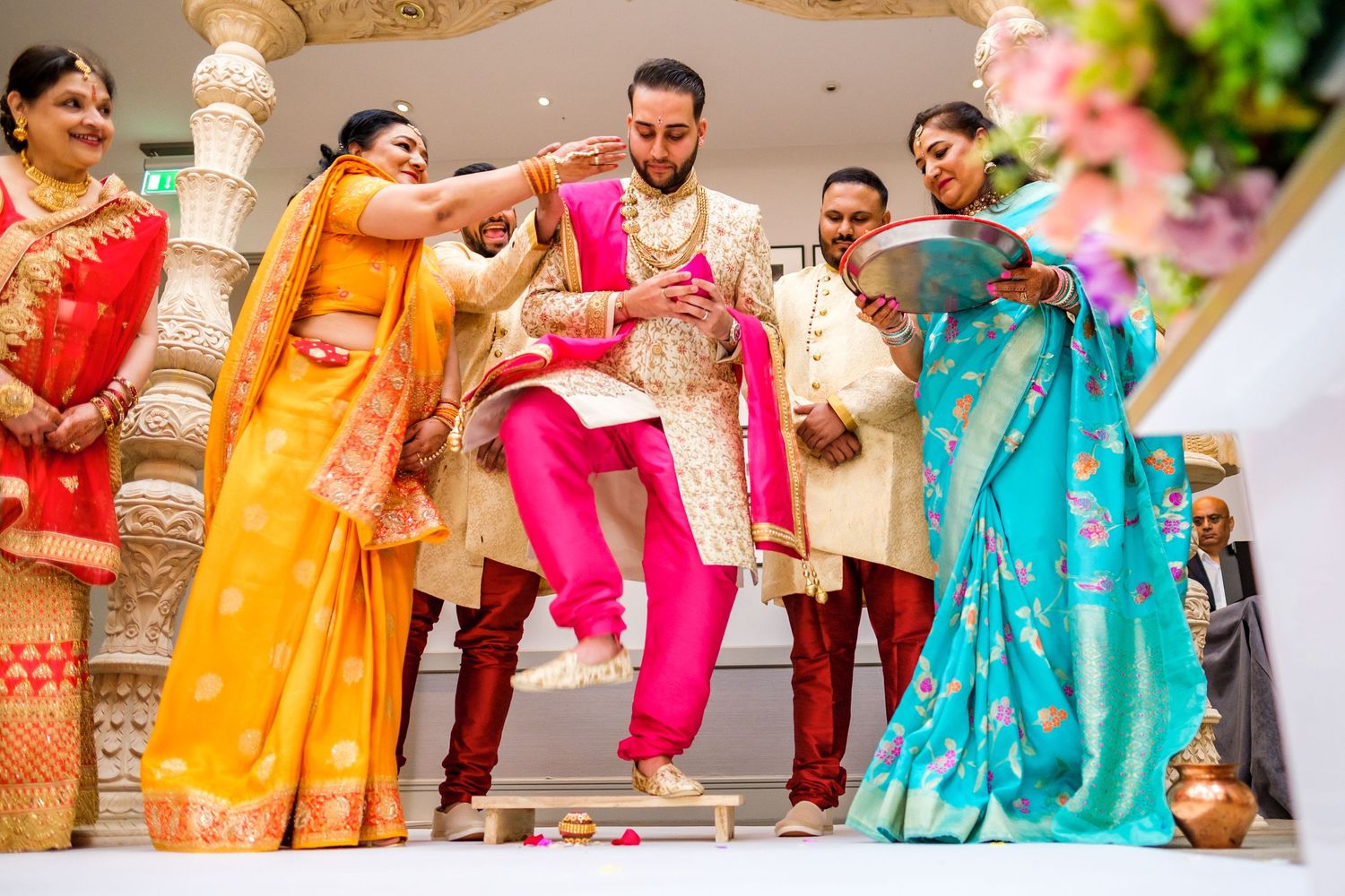 Wedding party members in vibrant saris and traditional outfits pose together for group photo.