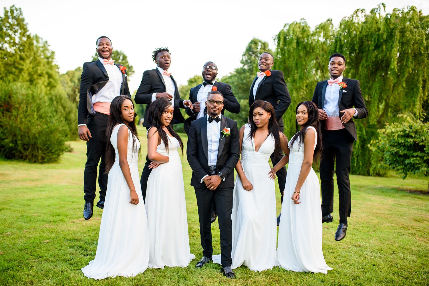 Wedding party poses playfully on lush green lawn with bridesmaids in white dresses and groomsmen in black tuxedos.