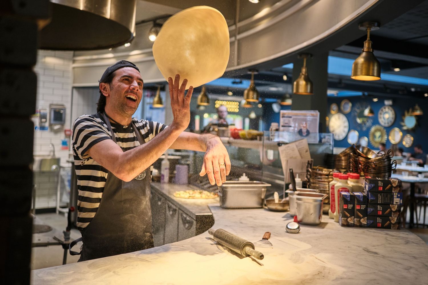 Chef in striped shirt tossing pizza dough in a restaurant kitchen with dramatic lighting.