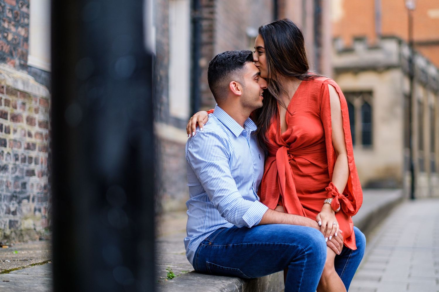Romantic couple in casual attire share intimate moments along a historic brick wall during an engagement photoshoot.