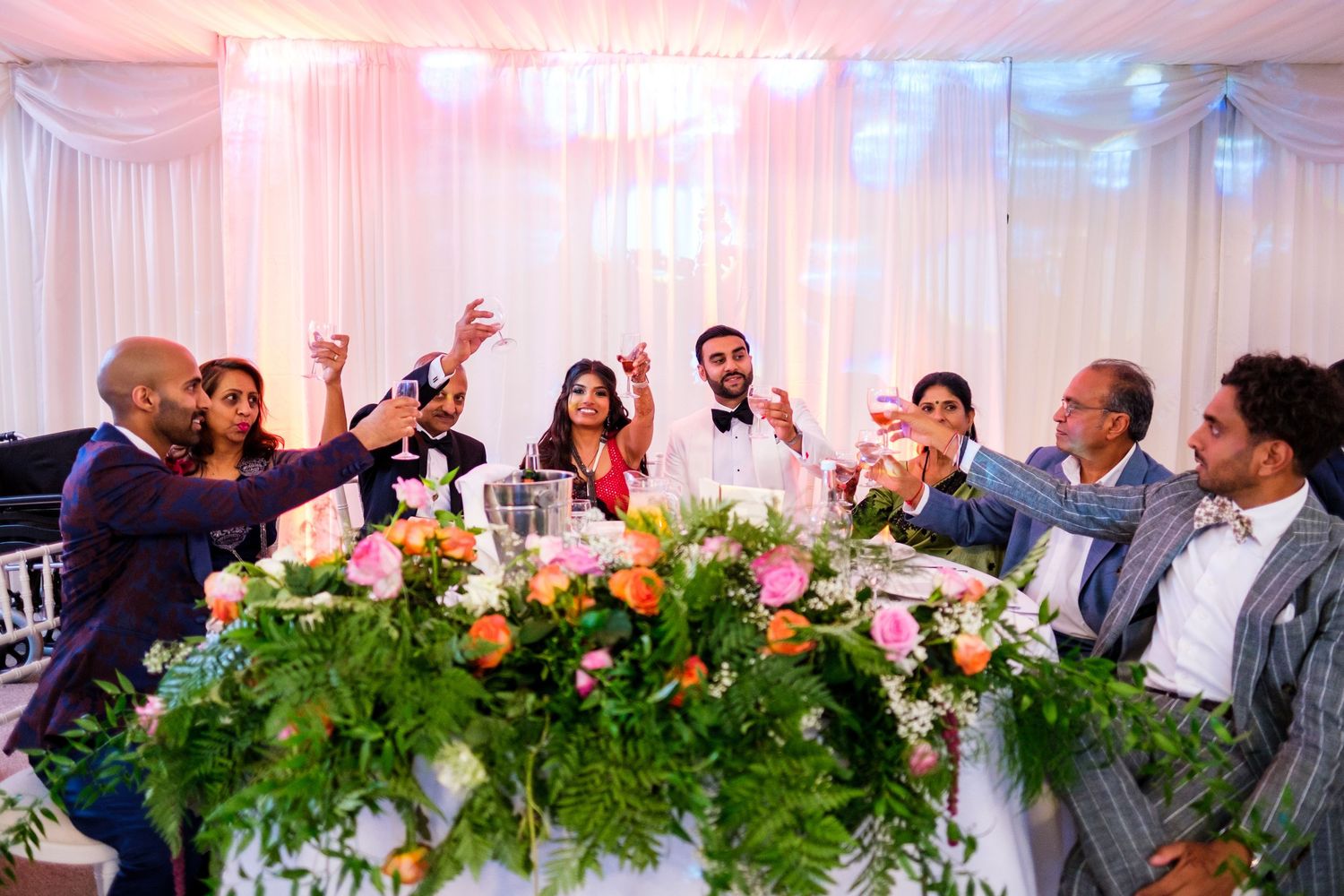 Wedding reception setup with pink lighting, white drapes and floral centerpieces on dining tables.