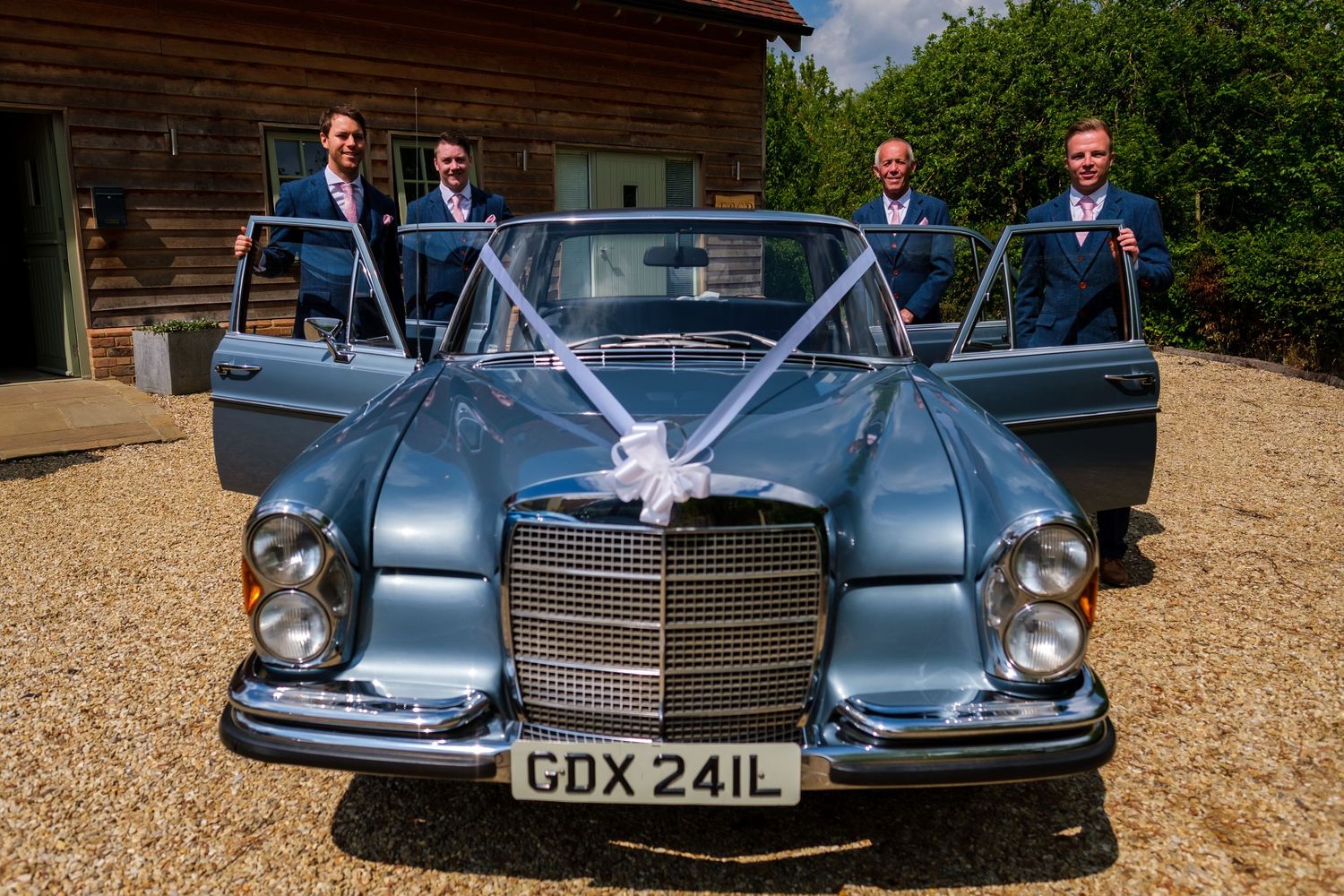 Classic vintage blue car parked on gravel driveway with elegant wedding party posing beside it.