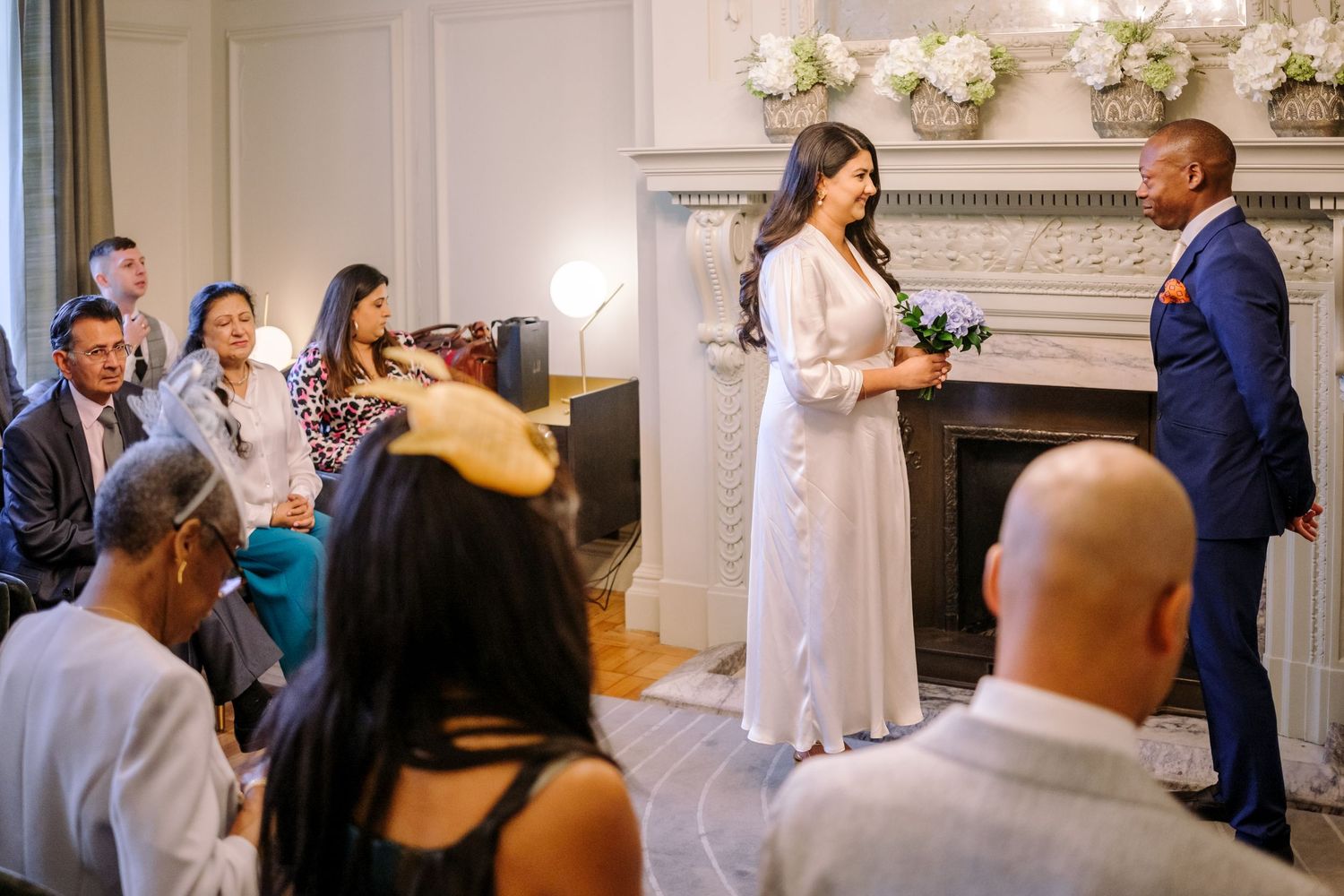 Wedding ceremony guests gather in an elegant indoor venue with white floral arrangements and classical decor.