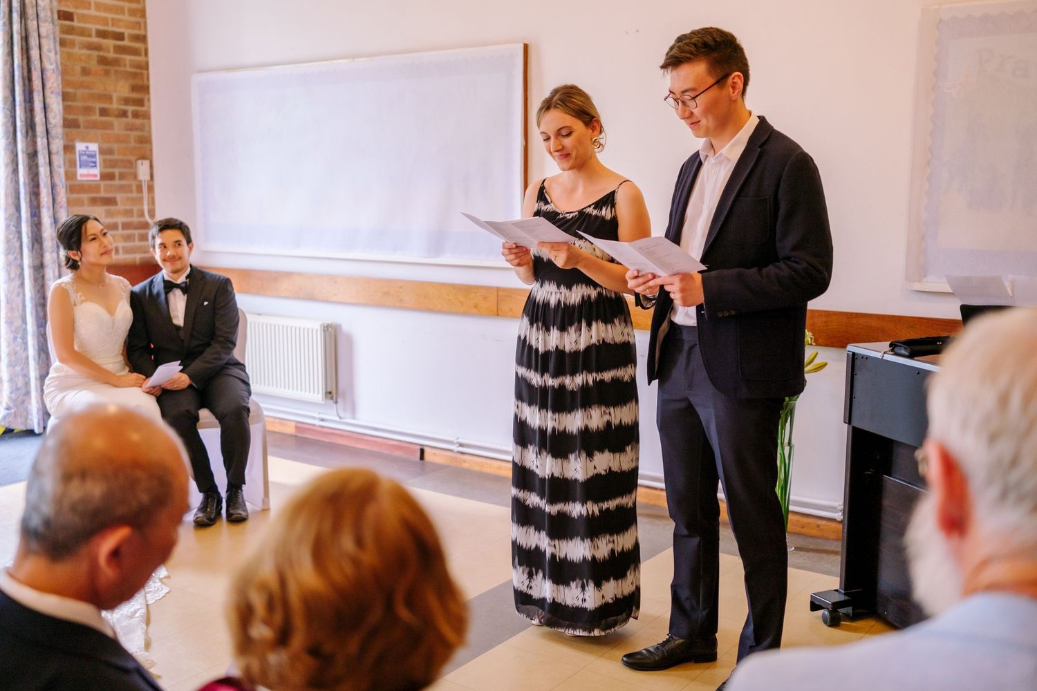 Wedding guests watch as speakers present at a ceremony in a bright indoor venue with brick walls.