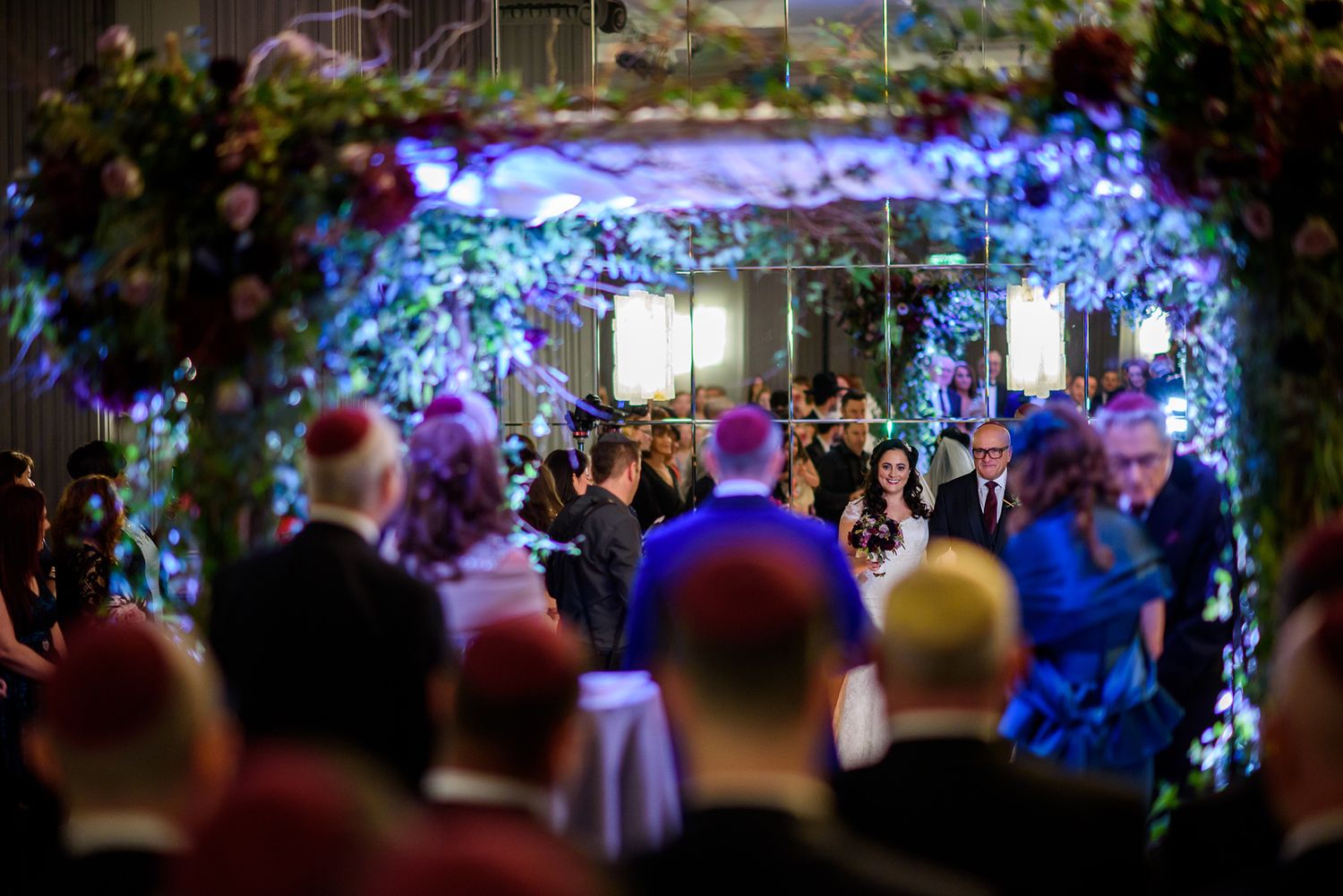 Wedding ceremony space with illuminated blue floral arch and crowd of guests in formal attire.